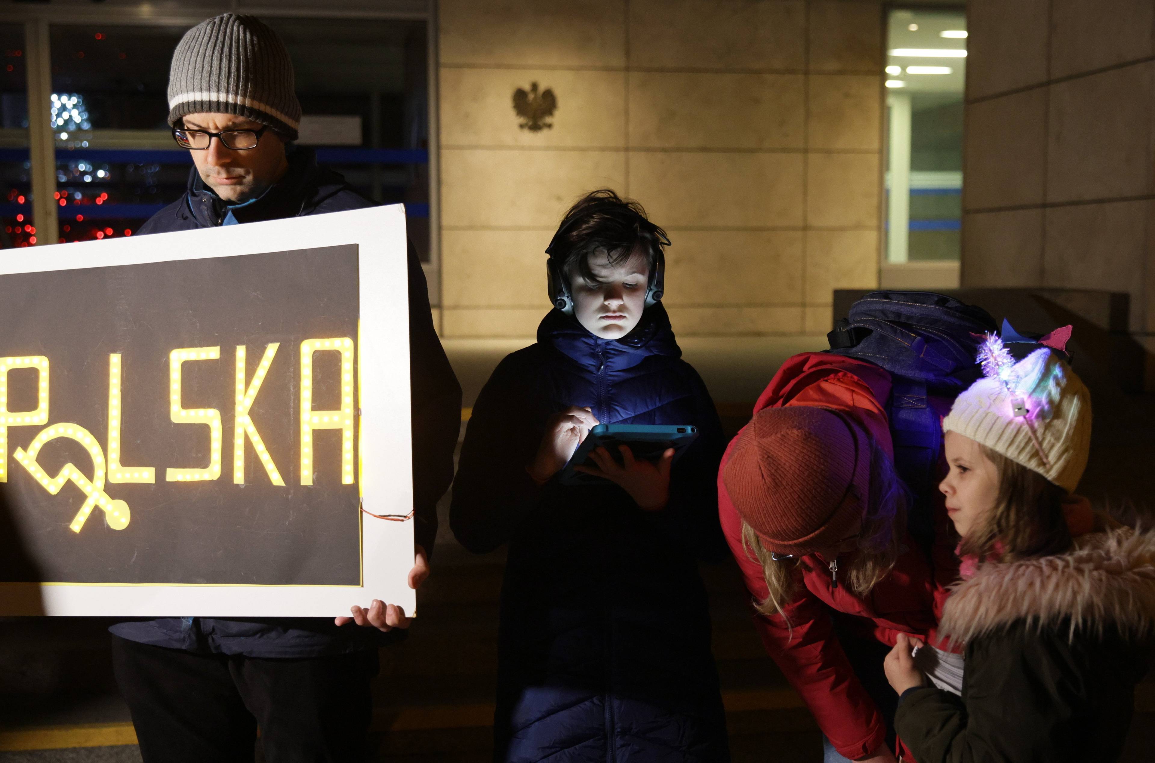 Grupa ludzi ze spuszczonymi głowami trzyma transparent POLSKA, w którym O jest zastapione znakiem wywróconego wózka inwalidzkiego