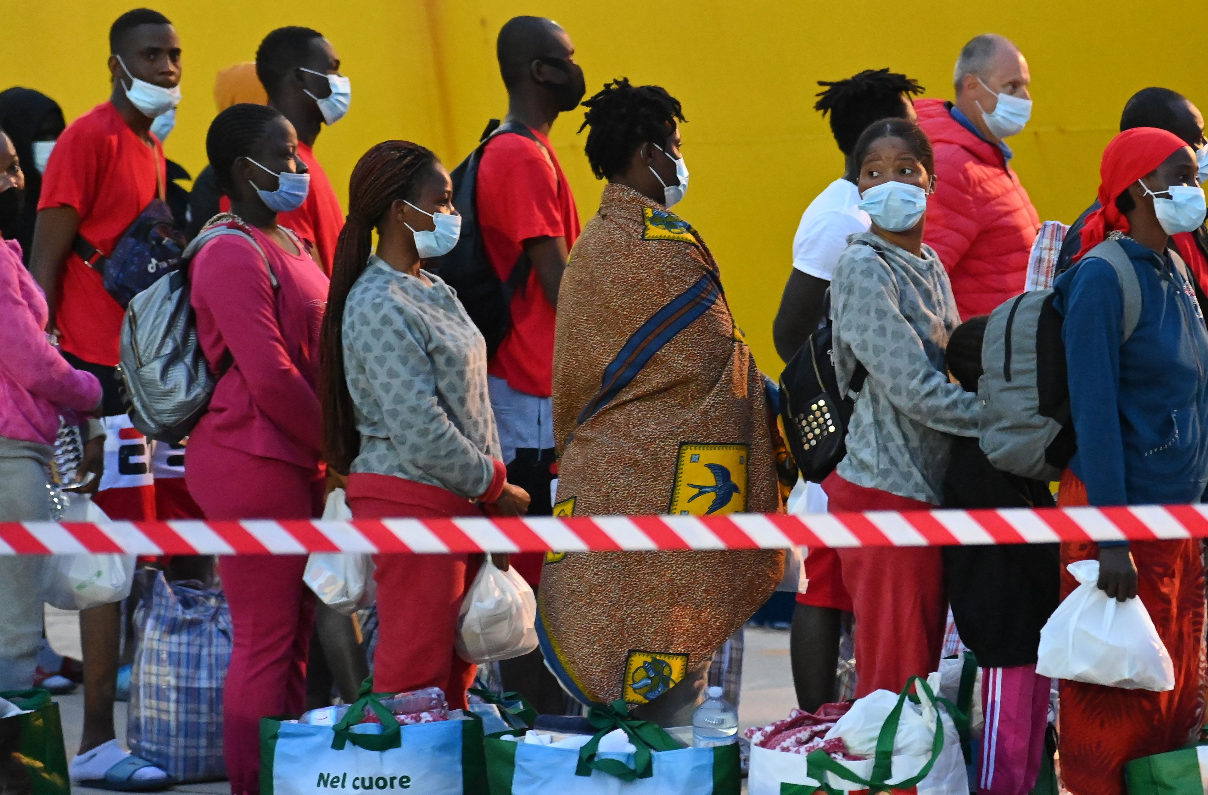 Kolejka migrantów z bagażami, oddzielona biało-czerwoną taśmą