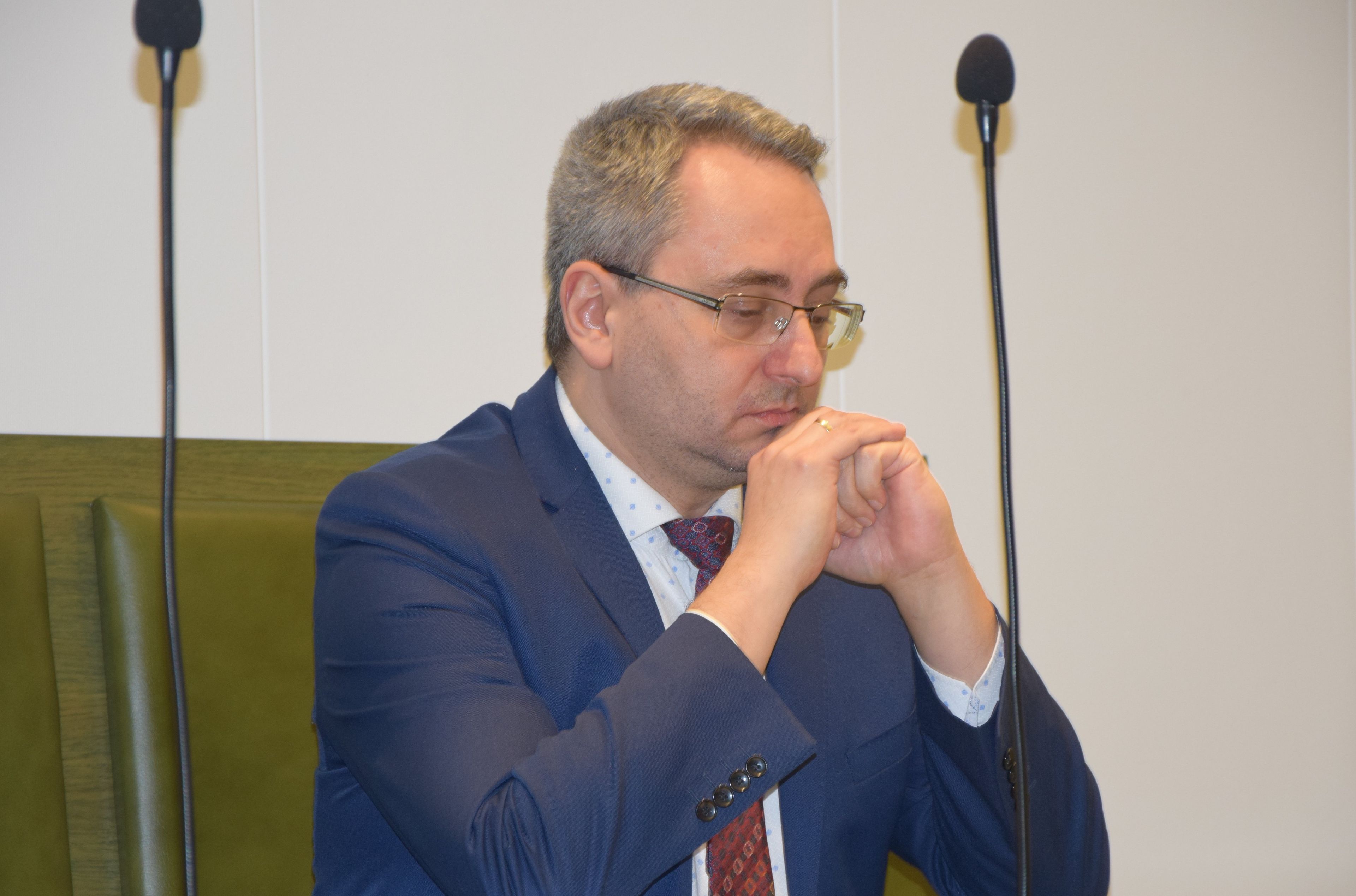 Mężczyzna w okularach siedzi za stołme w sali sądowej. Michał Lasota