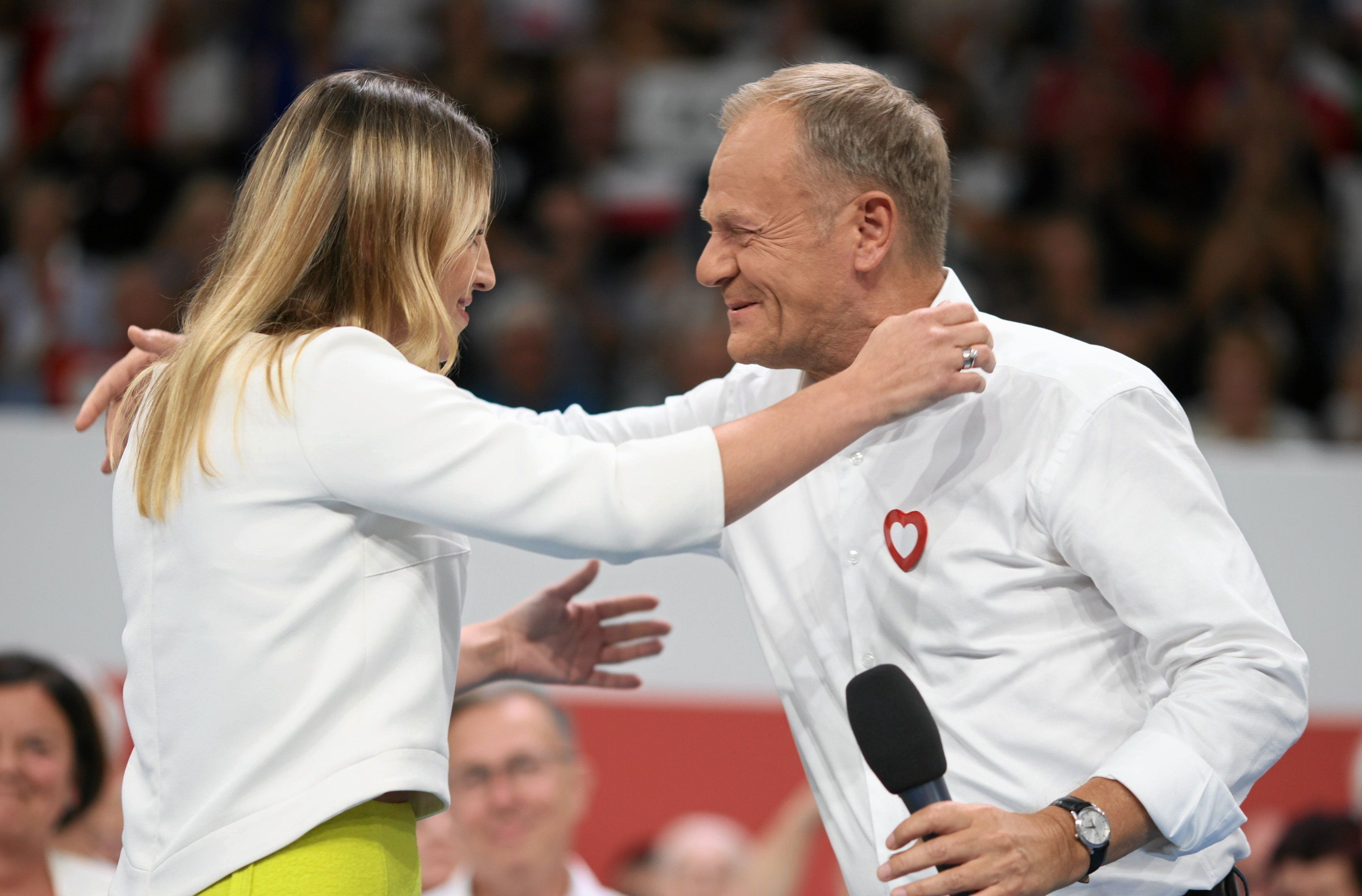 Kobieta z długimi włosami w białym żakiecie obejmuje w czasie publicznego wydarzenia mężczyznę w białej koszuli ze znakiem czerwonego serca