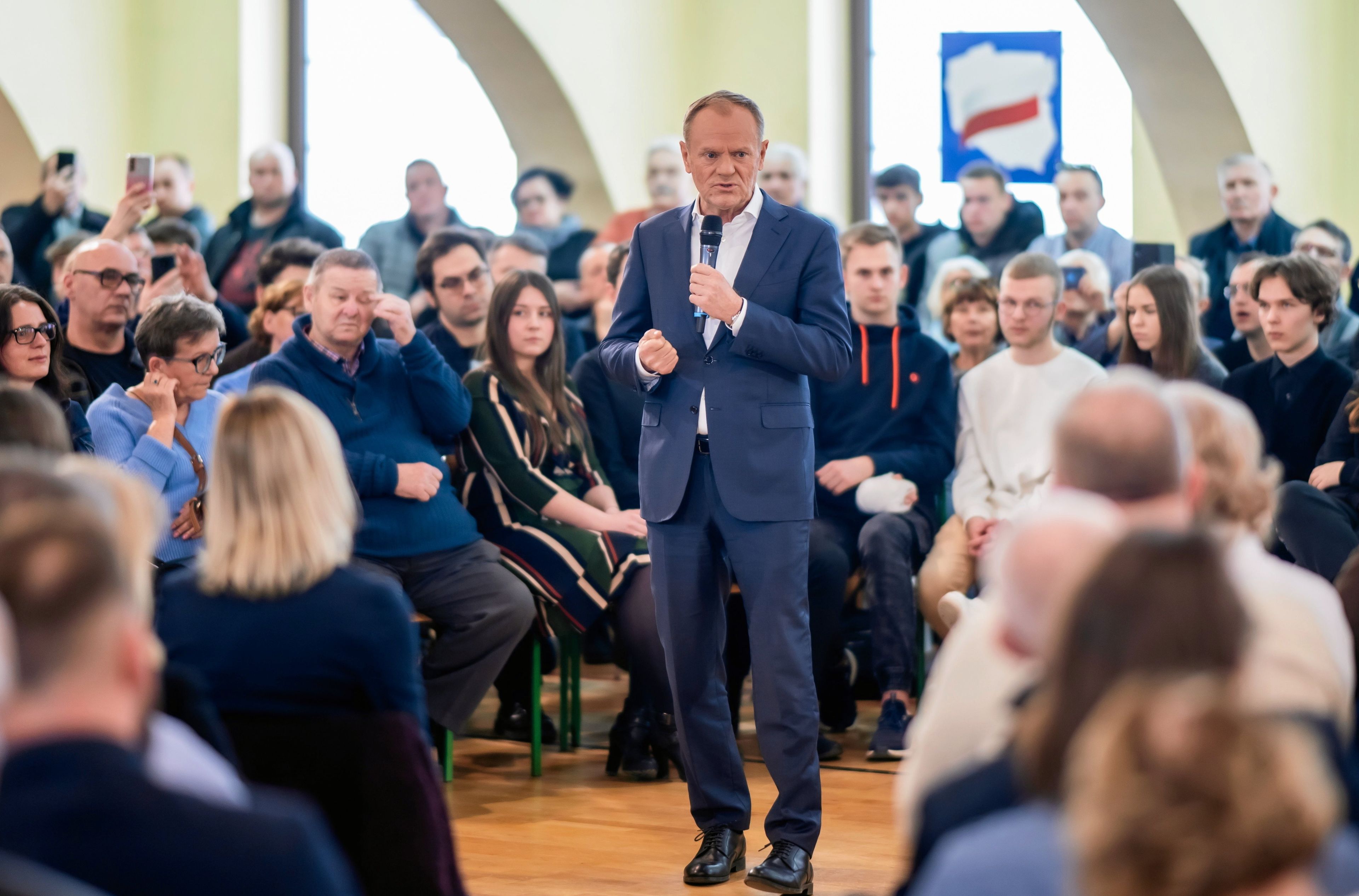 Donald Tusk przemawia stojąc wśród siedzących ludzi