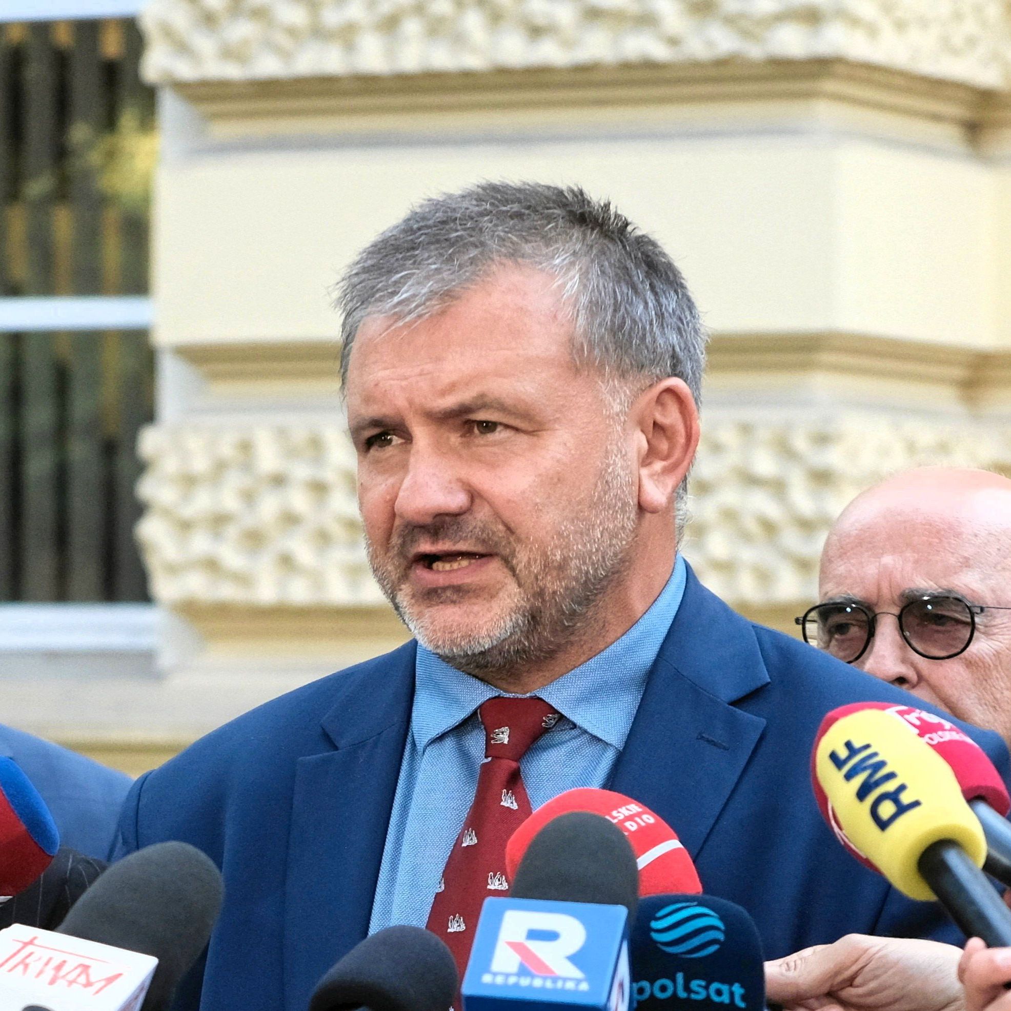 Krótko ostrzyżony mężczyzna w garniturzez odpowiada na pytania dziennikarzy