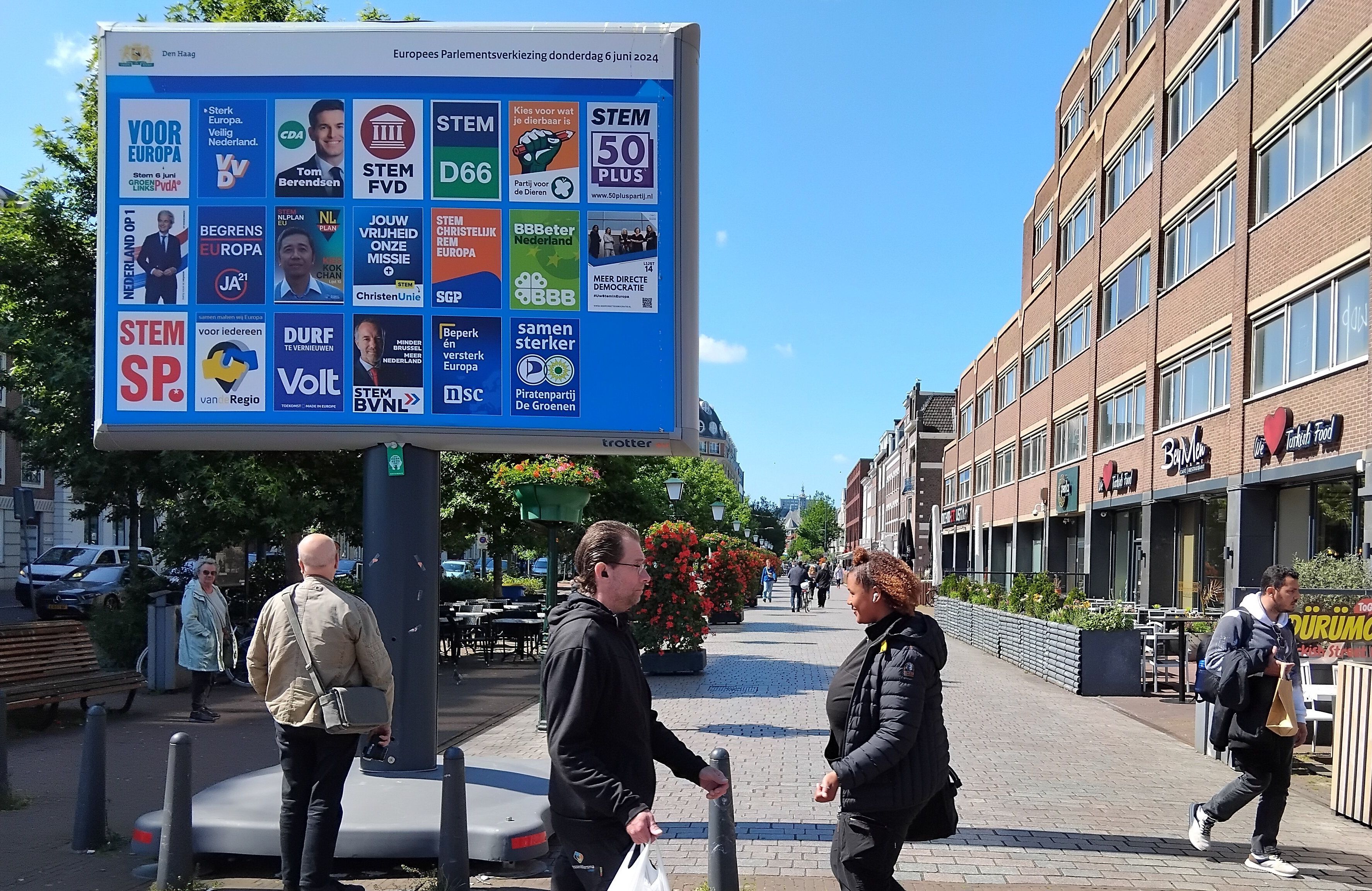 Widok na jedną z uliczek przy dworcu głównym w Hadze - z lewej strony przy chodniku stoi duży bilboard, a na nim 20 małych plakatów wyborczych każdej partii politycznej, która wystawia kandydatów w wyborach do Parlamentu Europejskiego 6 czerwca. Poza tym po ulicy chodzą ludzie, widać też ogródek restauracji z boku. Jest ładna, słoneczna pogoda, niebo niemal bez chmur.