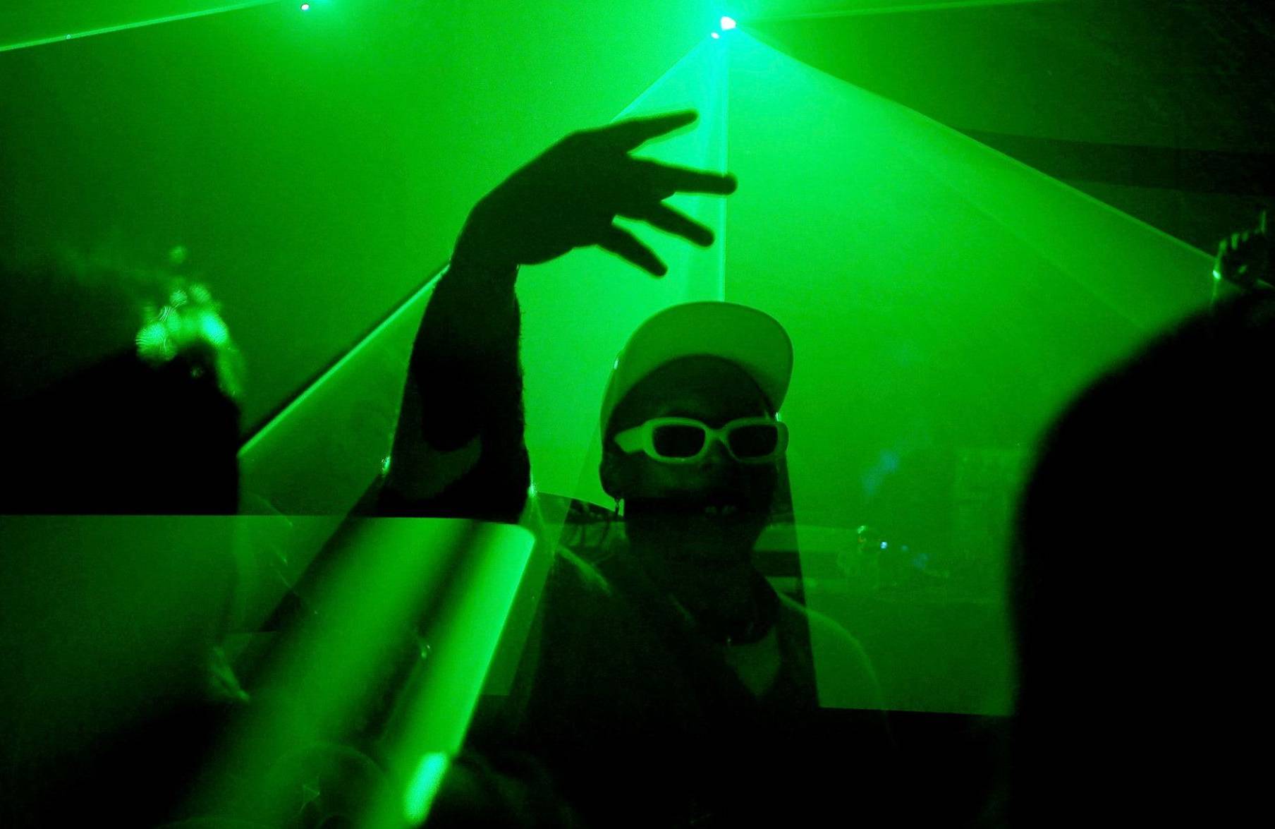 Mężczyzna w białej czapce i okularach przeciwsłonecznych tańczy na tle zielonego światła. Techno