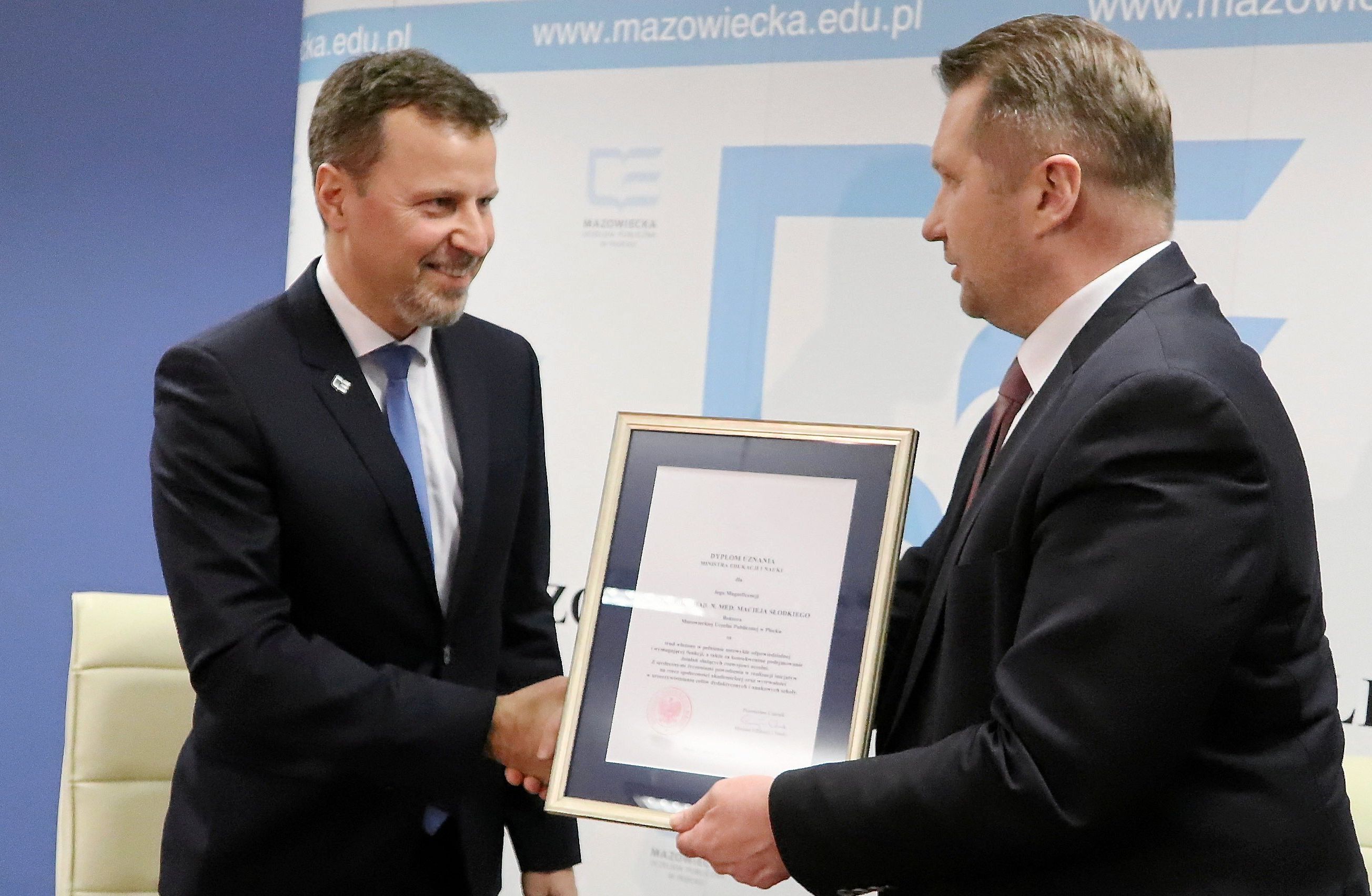 dwóch uśmiechniętych 40-latków w garniturach trzyma oprawiony dokument