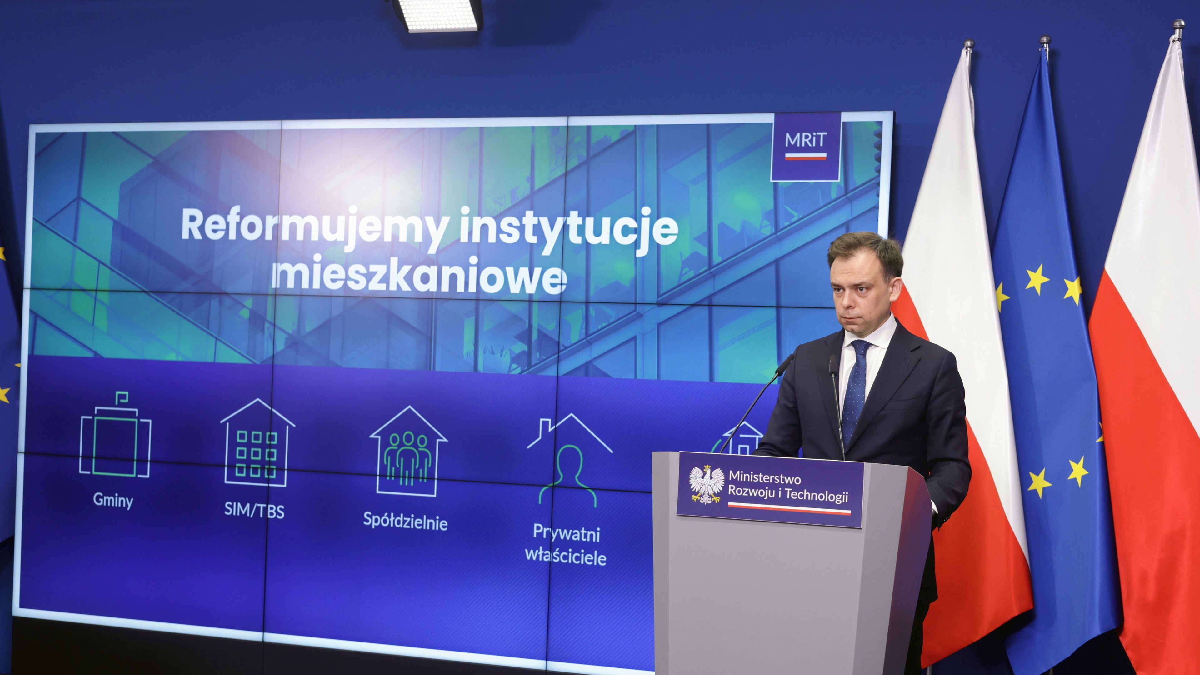 Mężczyzna - minister Domański - przemawia na tle wielkiego wyświetlonego na ekranie napisu Reformuje instytucje mieszkaniowe