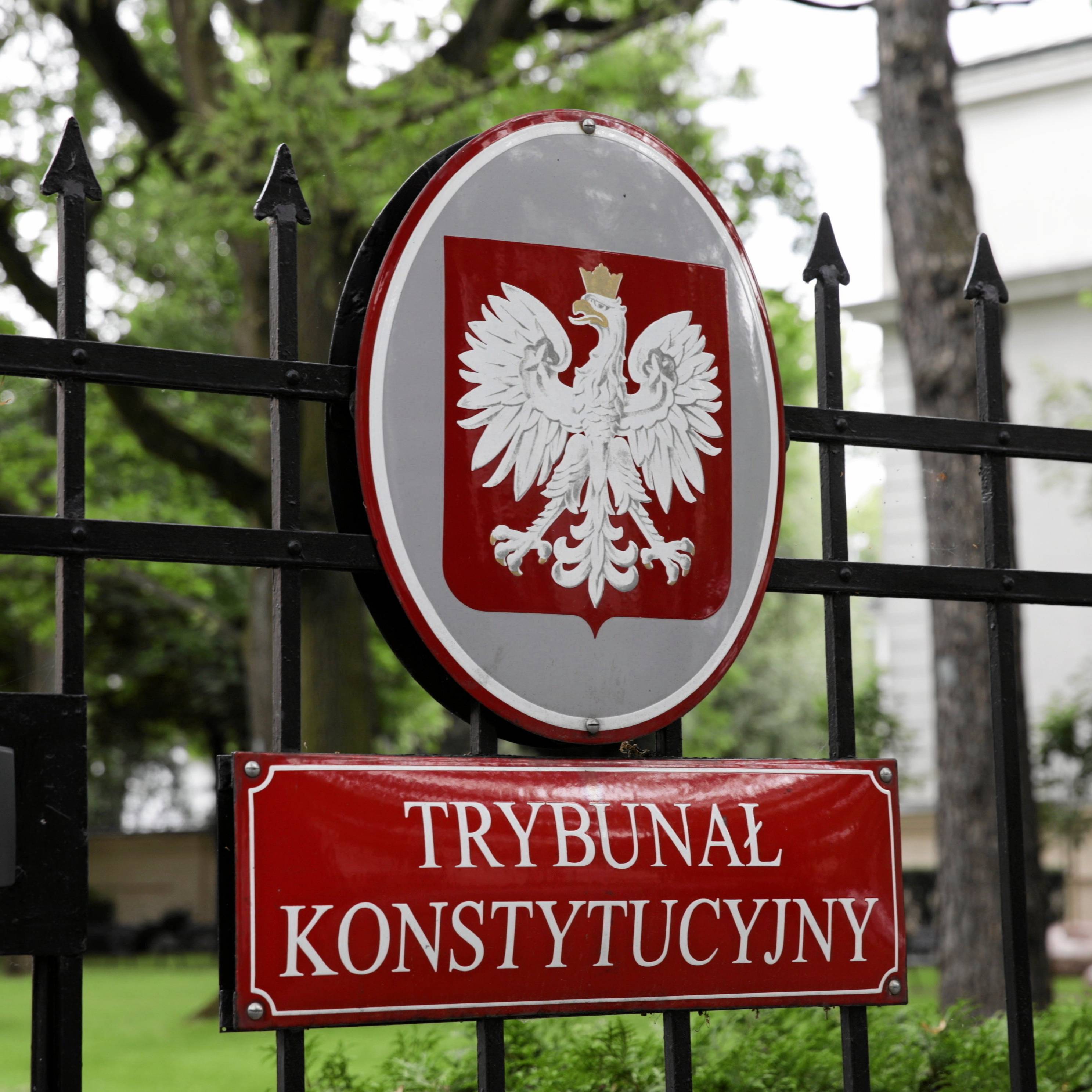 Zdjęcie na którym widać parkan otaczający siedzibę Trbunału Konstytucyjnego. Na płocie jest godło Polski i tabliczka z napisem Trybunał Konstytucyjny, za płotem jest bardzo zielono, dużo drzew, majaczy beżowy gmach