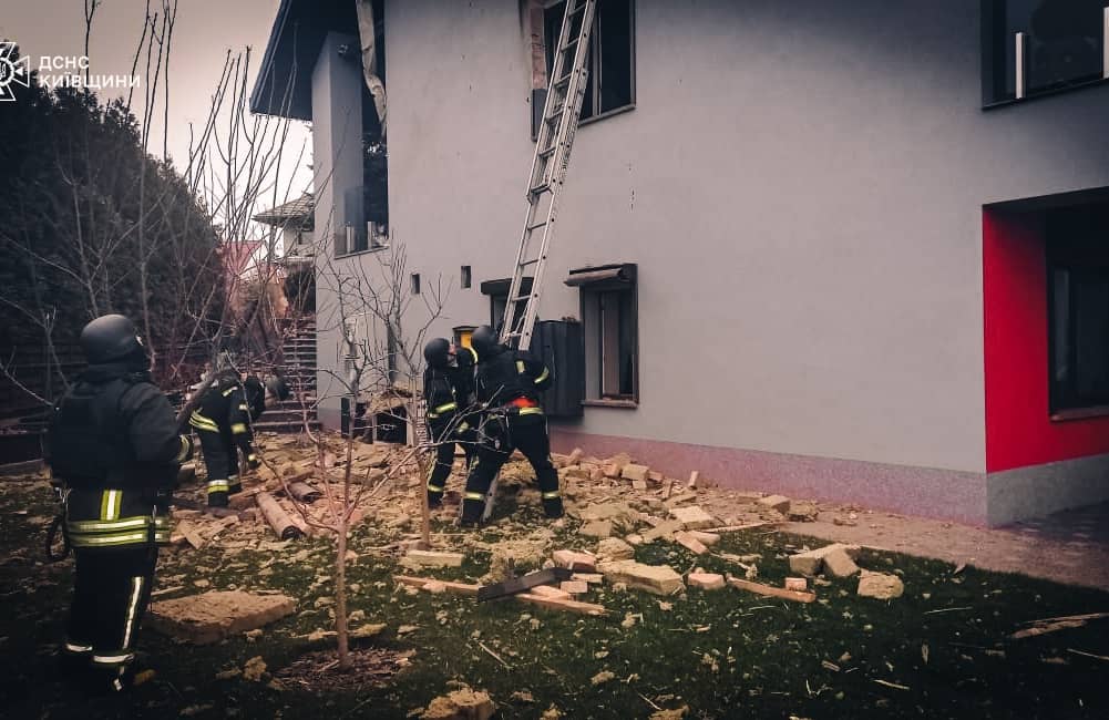 na zdjęciu jest czterech mężczyzn. Są ubrani w kombinezony ratowników o kolorze czarnym z żółtymi paskami odblaskowymi. Obok nich jest uszkodzony dom w szarym kolorze. Dwóch ratowników ustawia drabinę, próbując wspiąć się na dach domu.