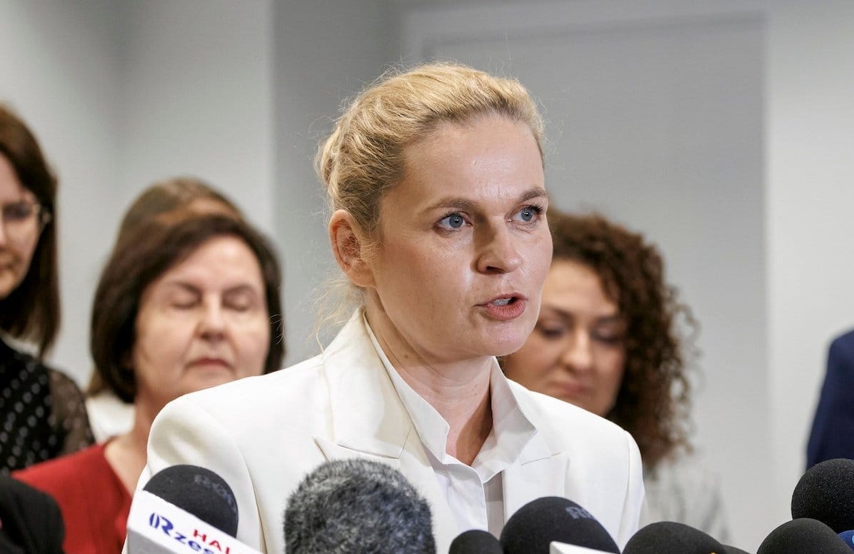 Ministra Edukacji Barbara Nowacka podczas konferencji prasowej. Ubrana w jasną marynarkę i koszulę, ma spięte z tyłu włosy