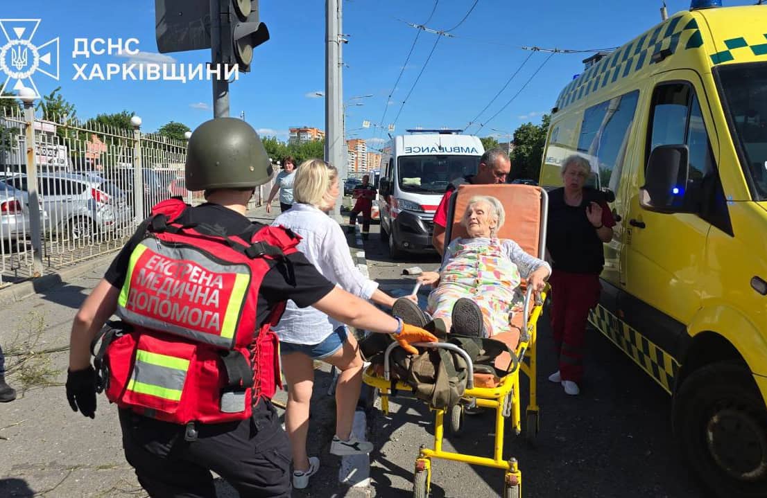 Starsza kobieta na noszach jest transportowana przez sanitariuszy w czerwonych kamizelkach i wojskowych hełmach do żółtego ambulansu