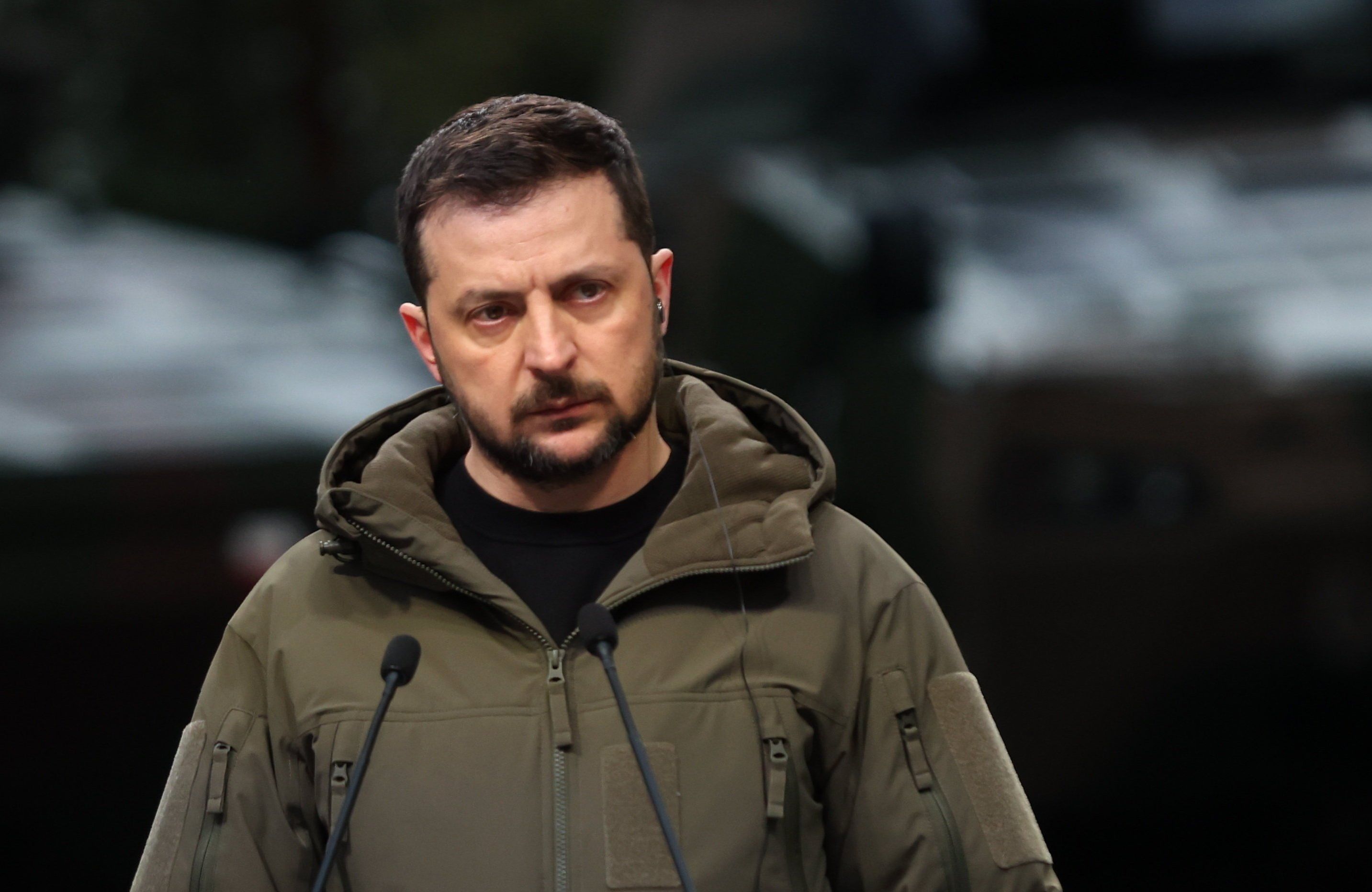 Wołodymyr Zełenski w zgniłozielonej kurtce podczas przemówienia na konferencji. Spogląda z groźna miną spod oka