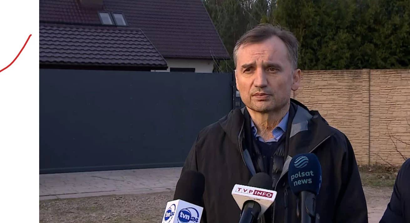 Mężczyzna z krótkimi ciemnymi włosami stoi przed ogrodzonym wysokim płotem domem i rozmawia z dziennikarzami