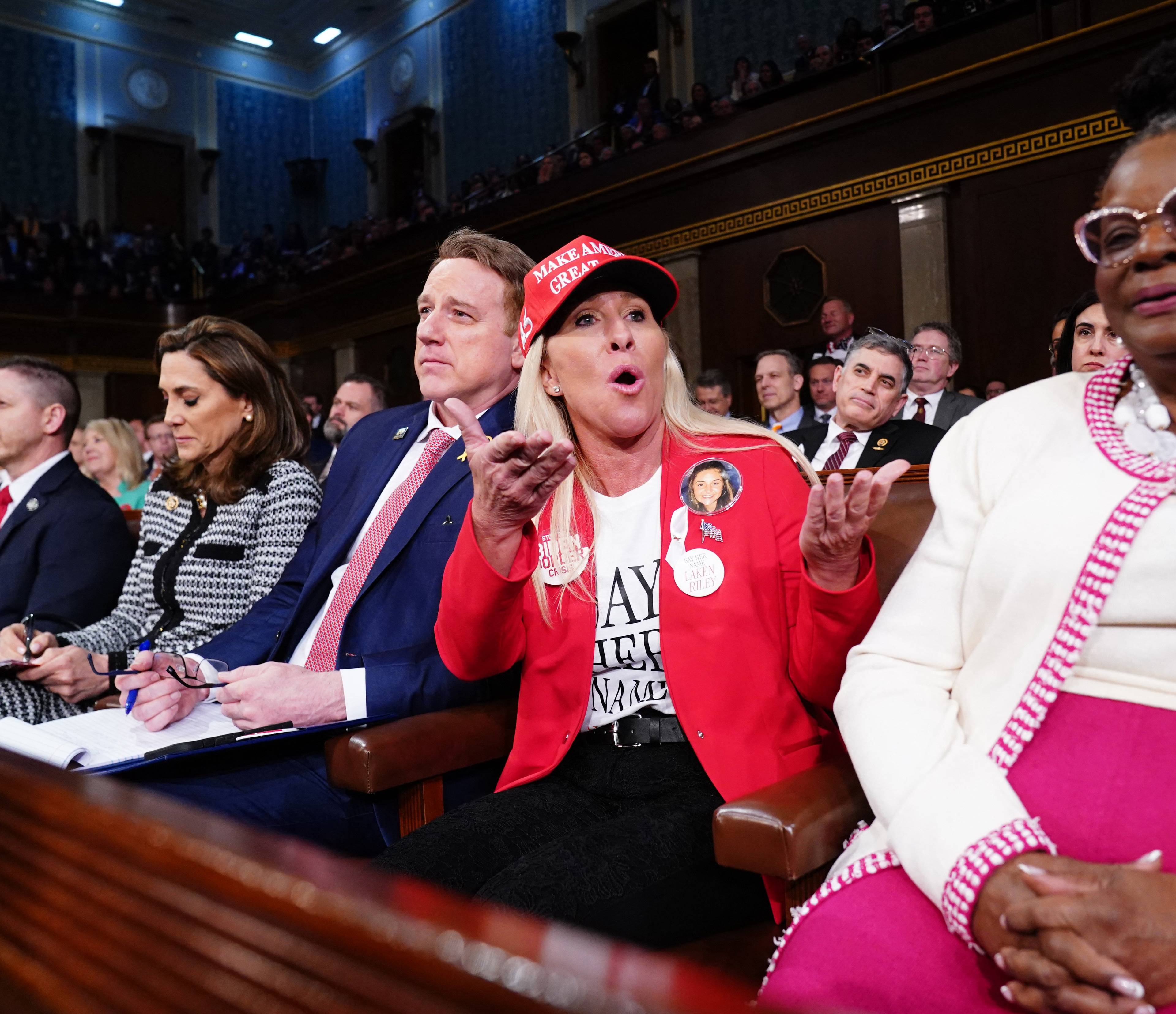 Kobieta ubrana na czerwono z czerwoną czapką na głowie krzyczy siedzie w ławach amerykańskiego kongresu