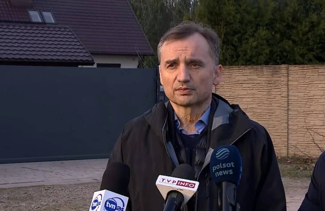 Szczupły mężczyzna w krótkich włosach stoi na tle domu otoczonego wysokim płotem rozmawiając z dziennikarzami