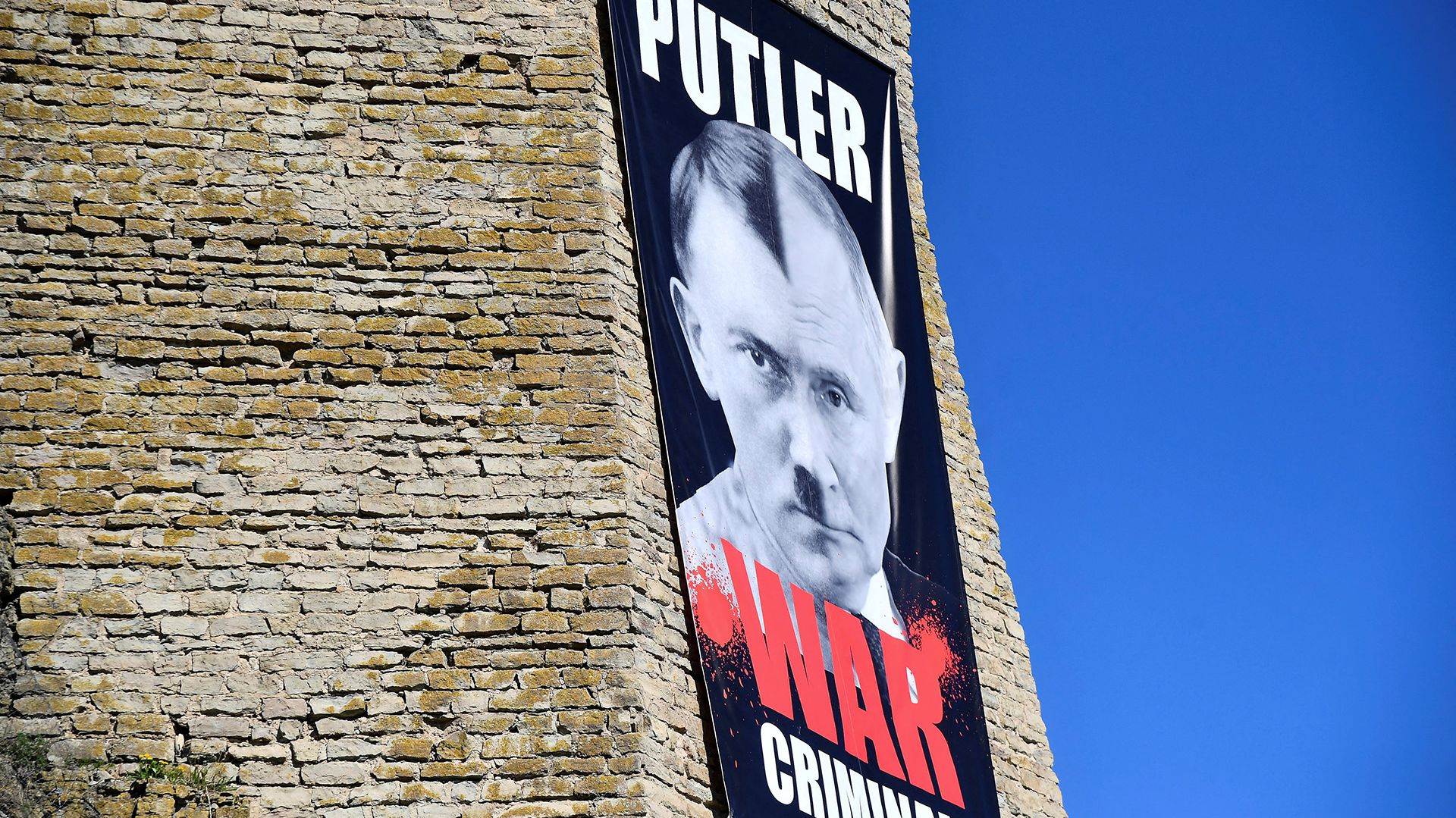 Zdjęcie przedstawia duży banner wywieszony na zamkowym murze, na bannerze połączone: pół twarzy Adolfa Hitlera i pół twarzy Władymira Putina, na bannerze napis: "Putler War Criminal"