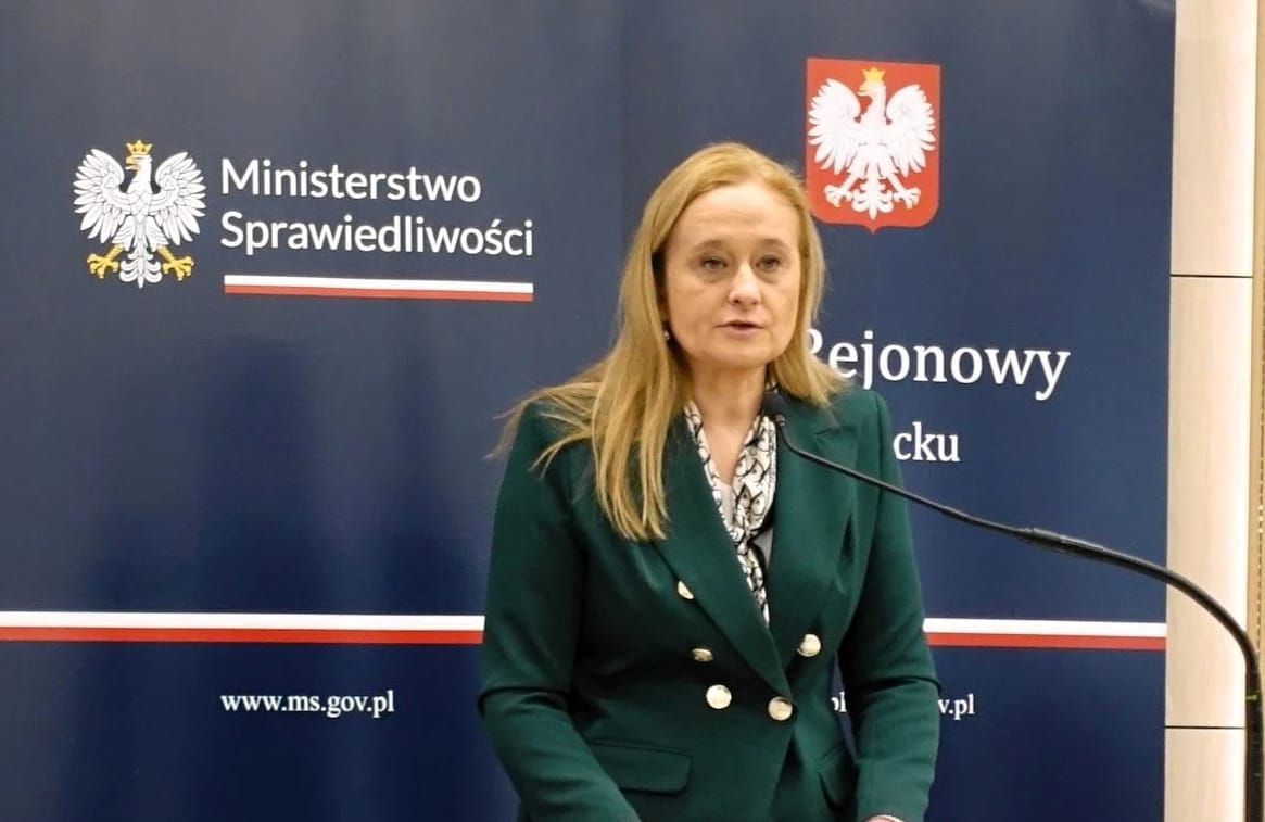 Kobieta, długie blond wóly porzemawia na tle logo ministerstwa sprawiedliwości