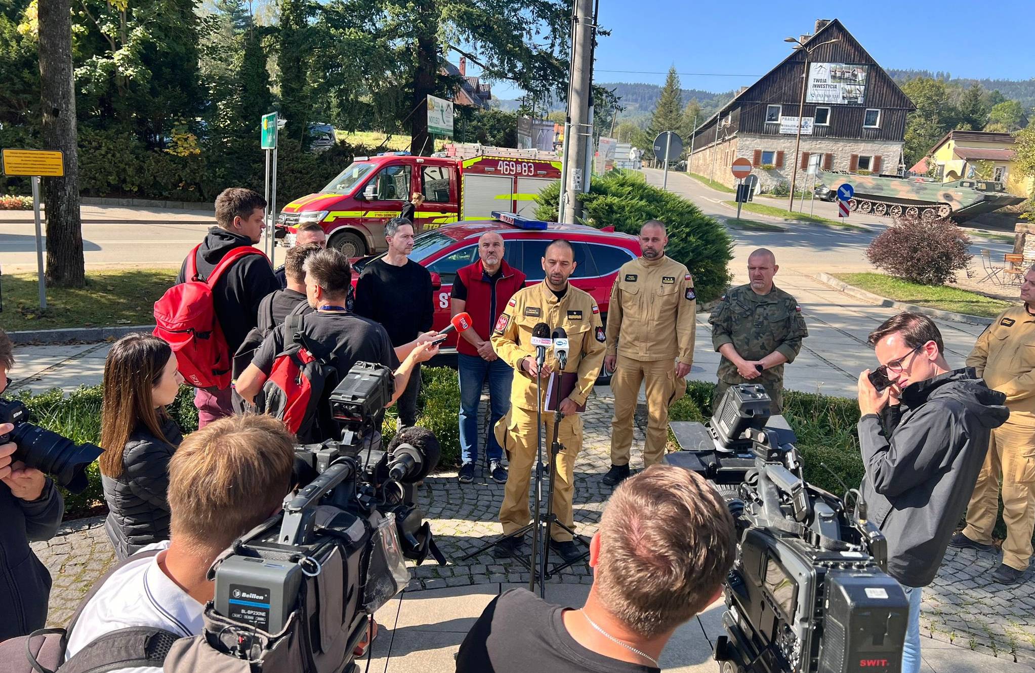 20.09.2024, briefing Państwowej Straży Pożarnej w Stroniu Śląskim. Strażak w żółtym kombinezonie przemawia przed mikforonem postawionym na statywie na ulicy. Wokół kamery telewizyjne i dziennikarze z mikrofonami.