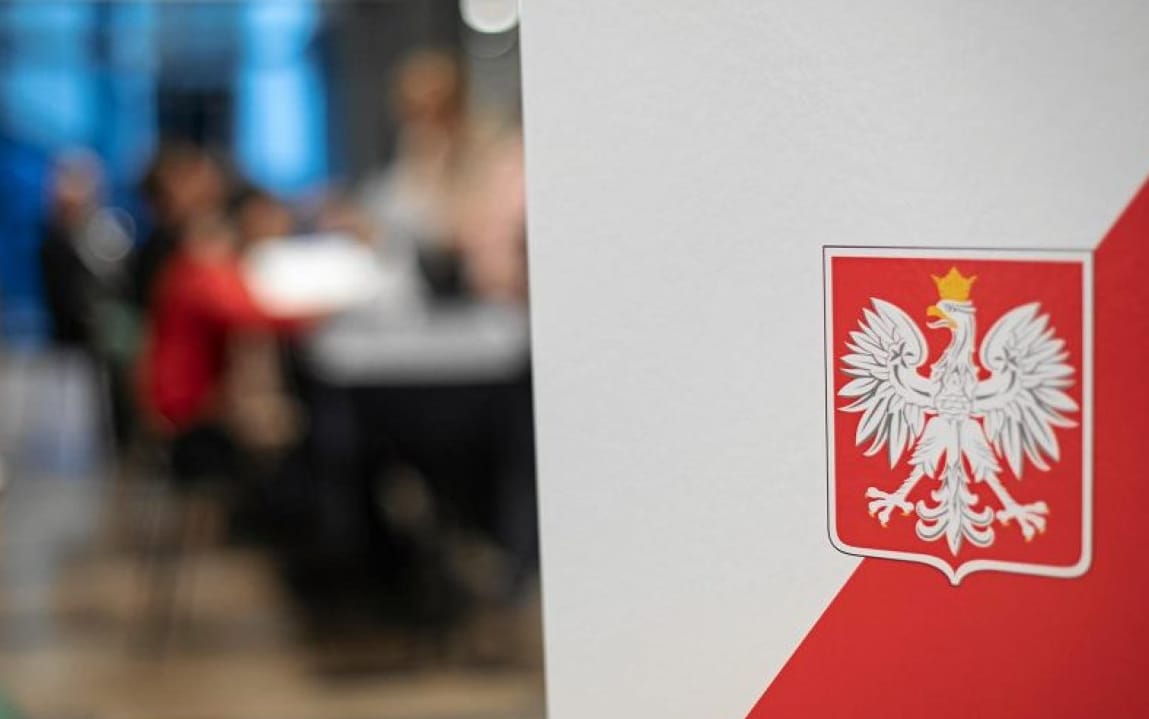 Urna wyborcza w biało-czerwonym kolorze, na pierwszym planie biały orzeł, godło Polski, w tle rozmazany obraz lokalu wyborczego