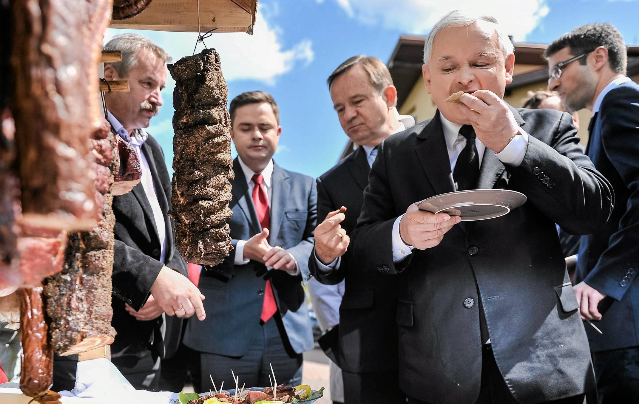 Jarosław Kaczyński stoi obok stoiska z mięsami, zjadając coś z talerzyka