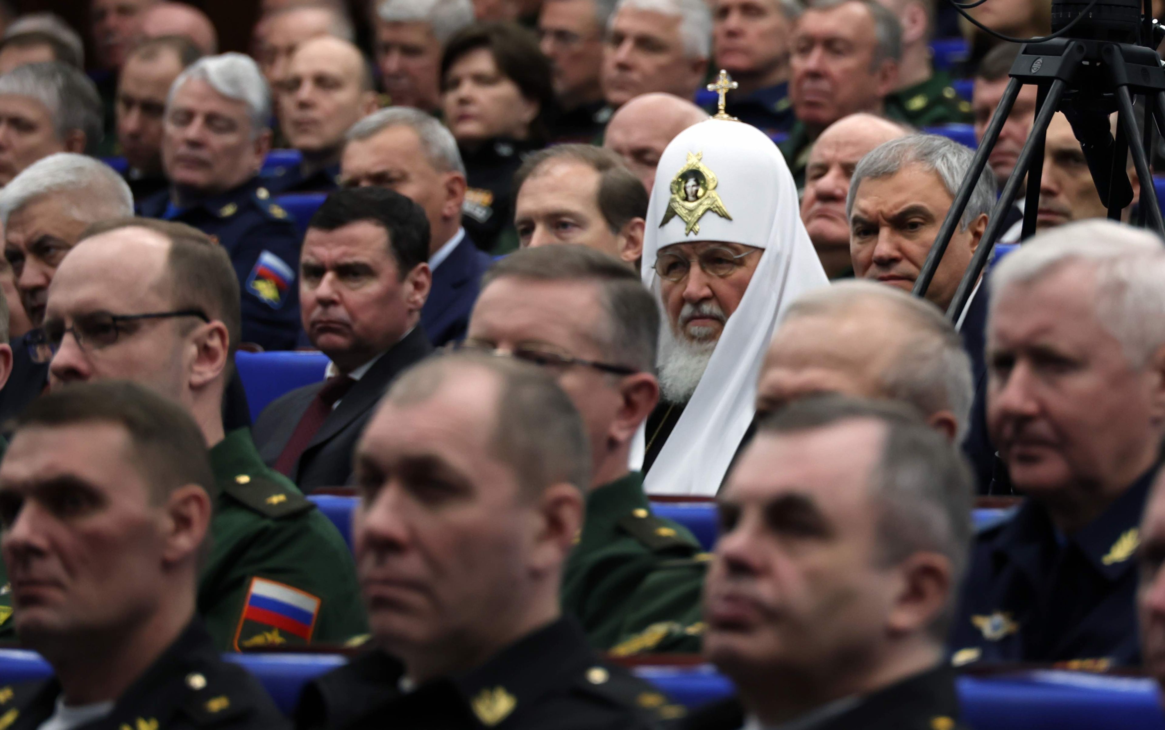 duchowny prawosławny a ceremonialnym białym nakryciu głowy z krzyżem siedzi na sali wśród wojskowych