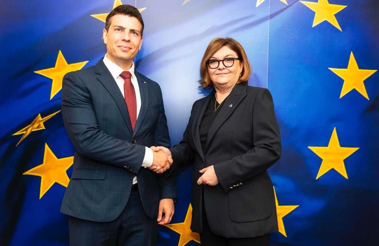 Na zdjęciu widać mężczyznę (po lewej) i kobiet (po prawej). Podają sobie dłoń. W tle widać flagę Unii Europejskiej