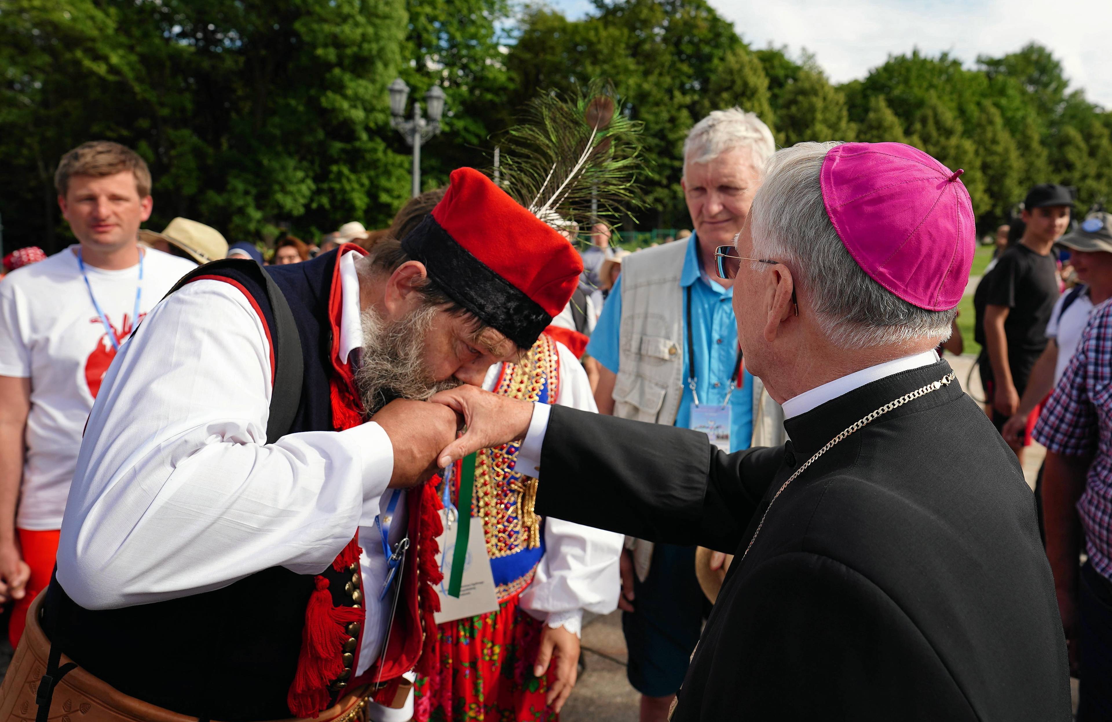 mężczyzna w krakowskim stroju, z pawim piórem przy czapce całuje w rękę biskupa w czarnej sutannie i purpurowej piusce