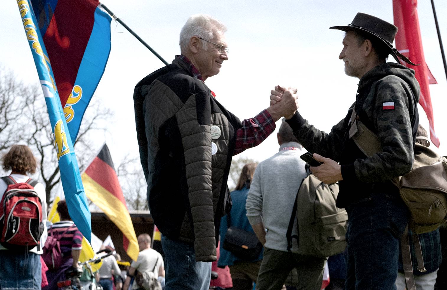 Dwóch mężczyzn trzyma dłonie w przyjacielskim uścisku, jeden z nich trzyma flagę, na pierwszym planie osoba leży na trawie, w tle tłum osób
