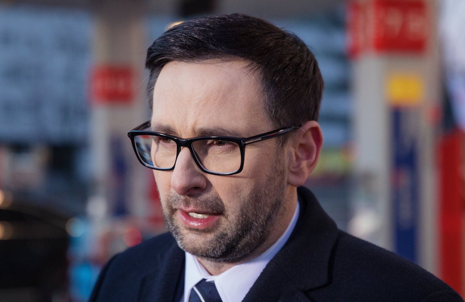 Daniel Obajtek, prezes Orlenu, na tle stacji benzynowej Orlen. Ma na sobie płaszcz i granatowy krawat w jasne pasy. Nosi okulary w ciemnych, prostokątnych oprawkach.