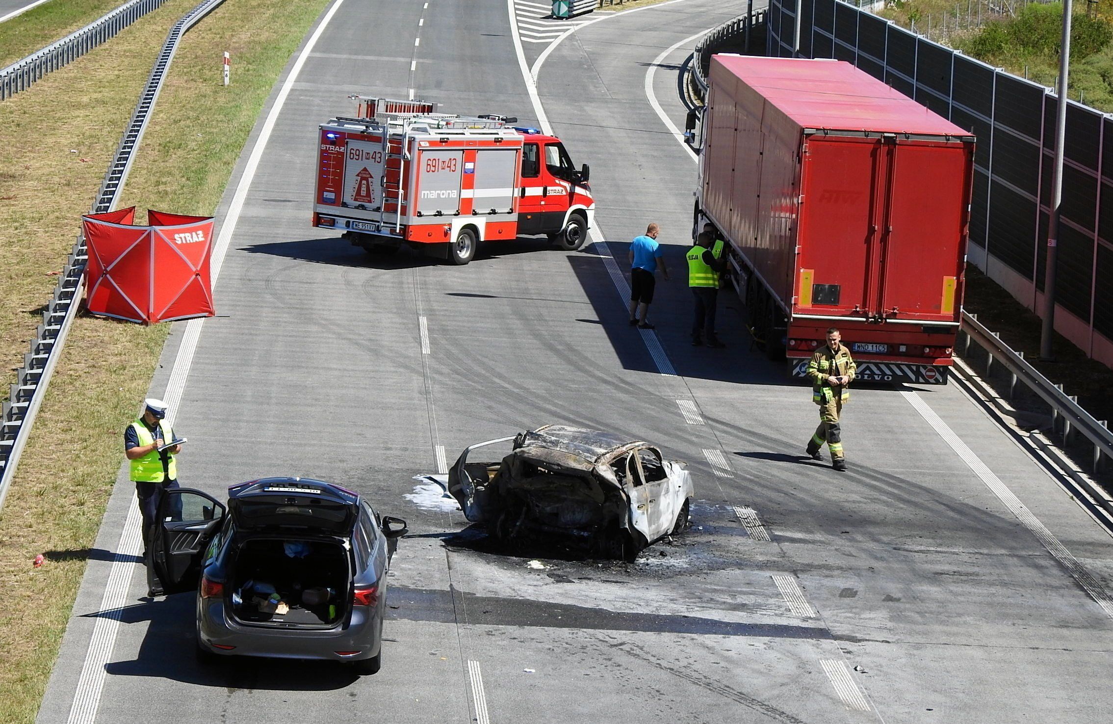 wypadek na trasie szybkiego ruchu, na prawym pasie spalony wrak samochodu, po lewej stronie czarne auto z otwartymi drzwiami, policjant robiący zdjęcia, w oddali wóz straży pożarnej