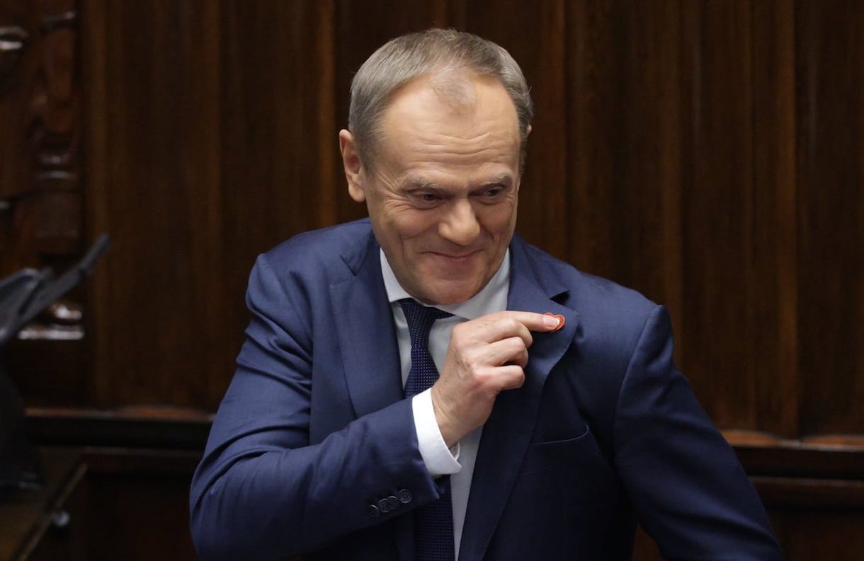 Donald Tusk na sejmowej mównicy uśmiecha się i wskazuje palcem biało-czerwone serduszko, które ma wpięte w klapę marynarki.