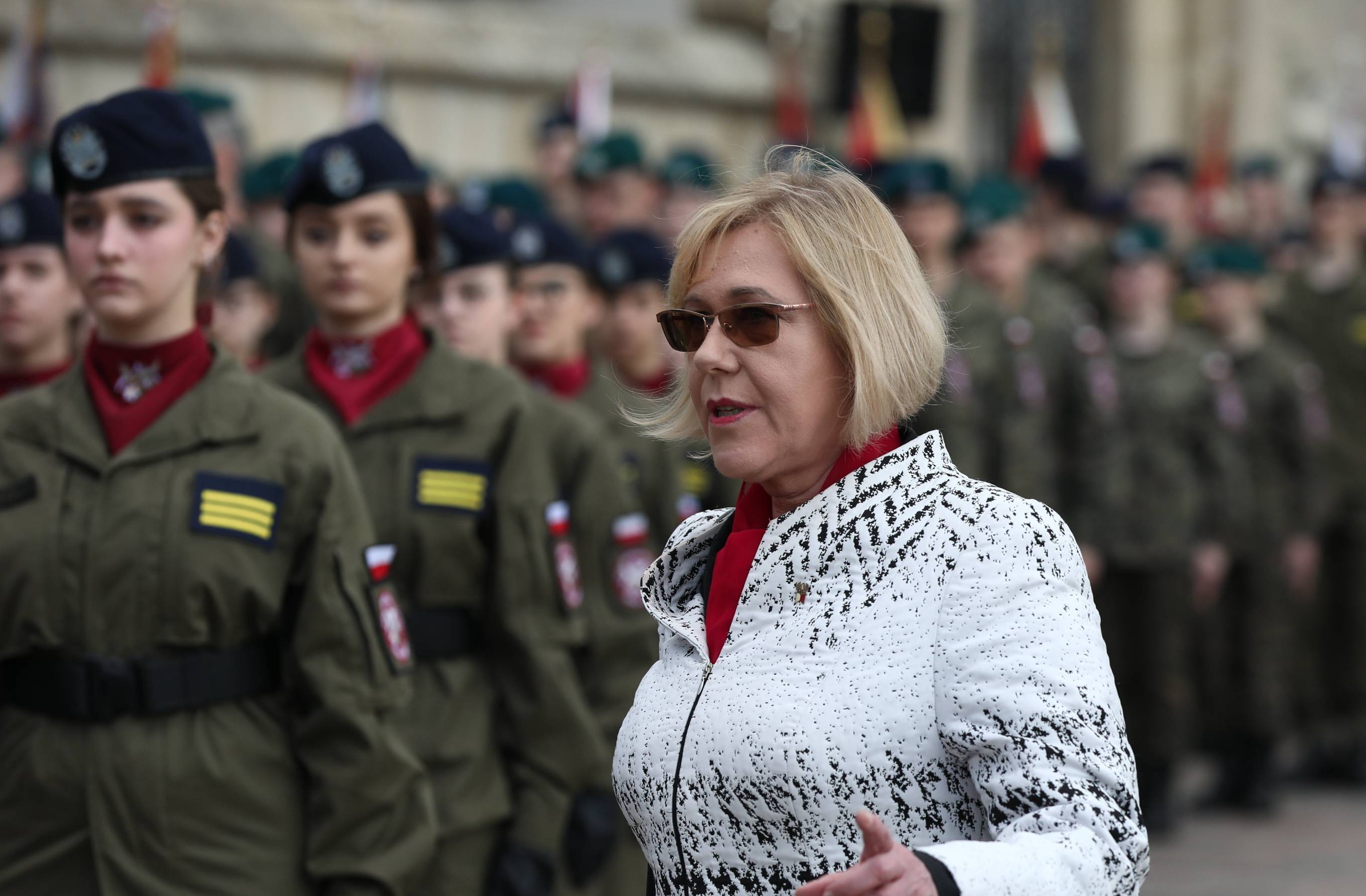 Kobieta w jasnej kurtce i ciemnych okularach stoi obok młodzieży w wojskowych mundurach