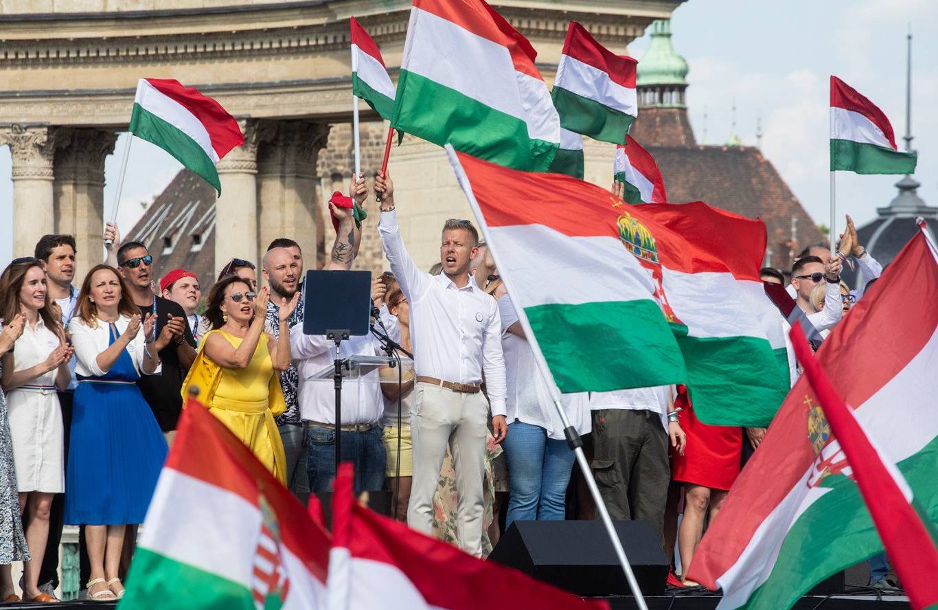 Mężczyzna w białej koszuli stoi na scenie, trzymając węgierską flagę