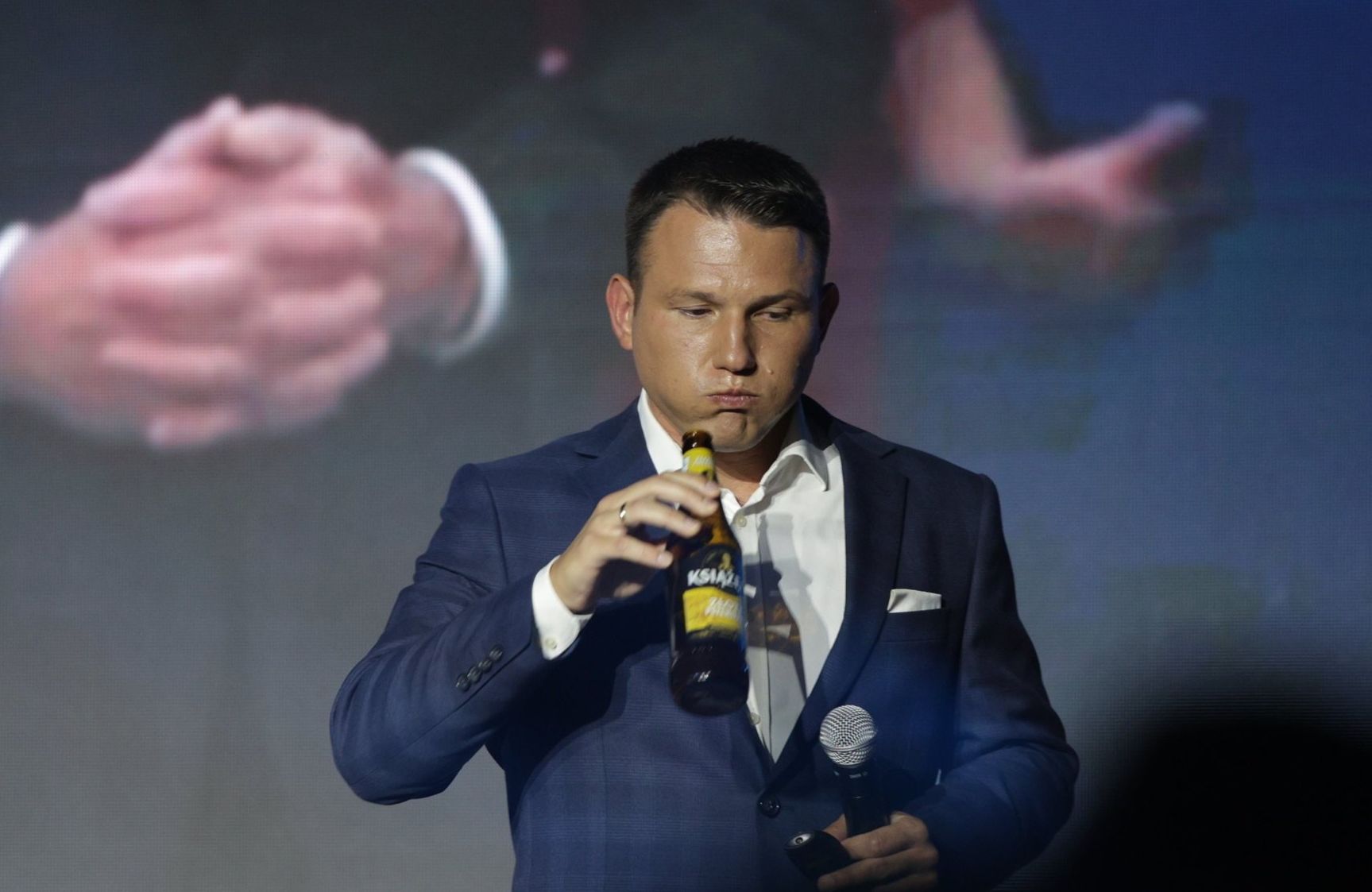 Sławomir Mentzen w garniturze stoi na scenie, w dłoniach ma mikrofon i butelkę piwa