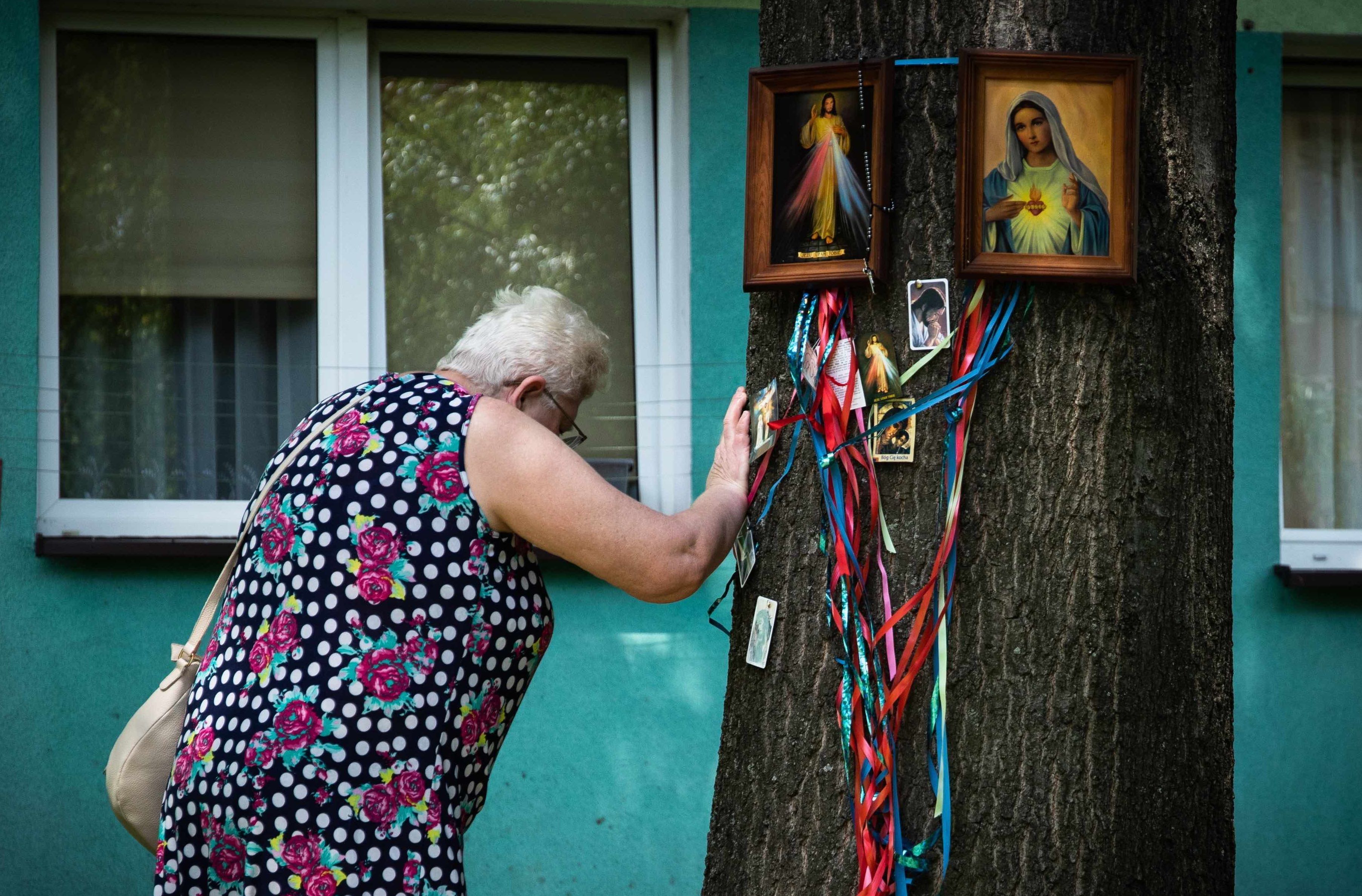 Kobieta opiera rękę o drzewo, na którym wiszą obrazki święte i wstążki. W tle widać okna bloku mieszkalnego.