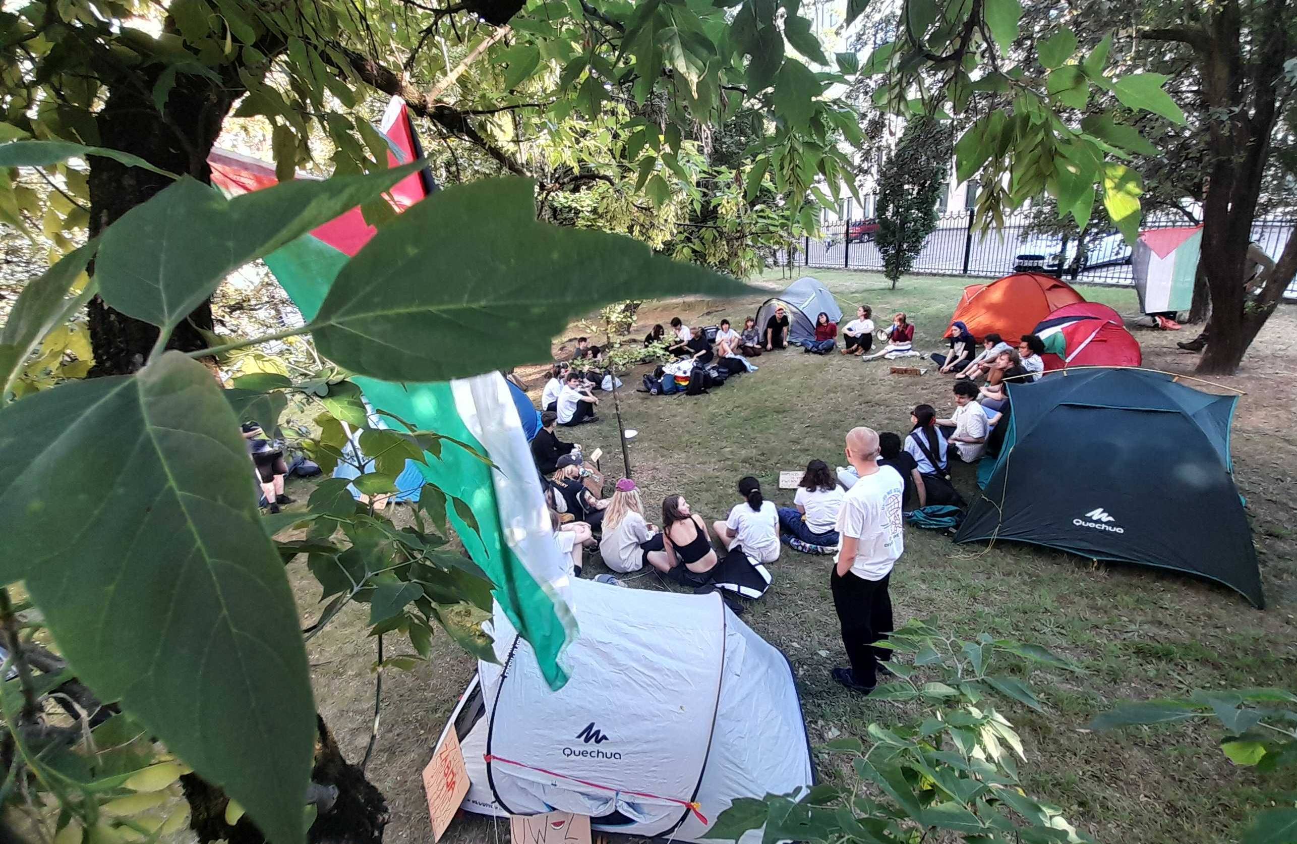 Grupa studentów siedzi na trawie w kółku. Wokół nich namioty, w których będą spać podczas okupacji terenu Uniwersytetu Warszawskiego