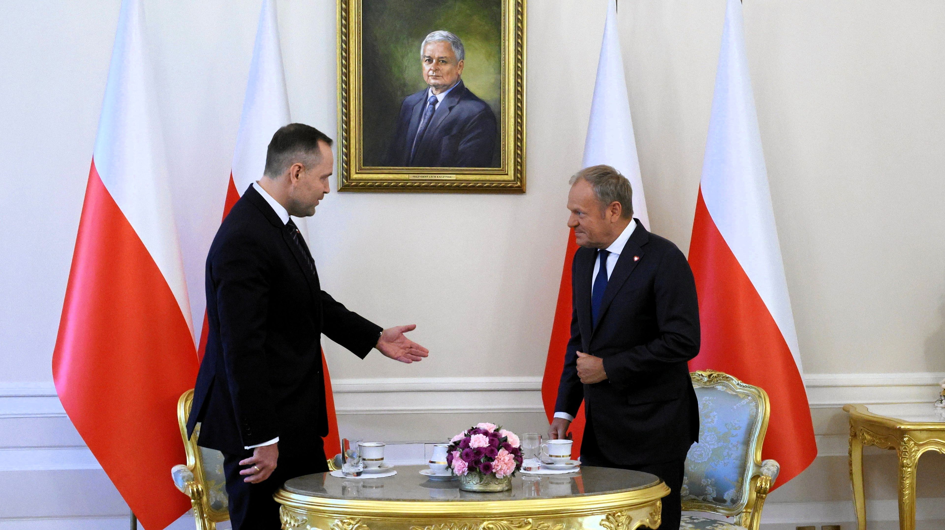 Dwaj mężczyźni stoją przy okrągłym stopliku, jeden gestem zaprasza drugiego