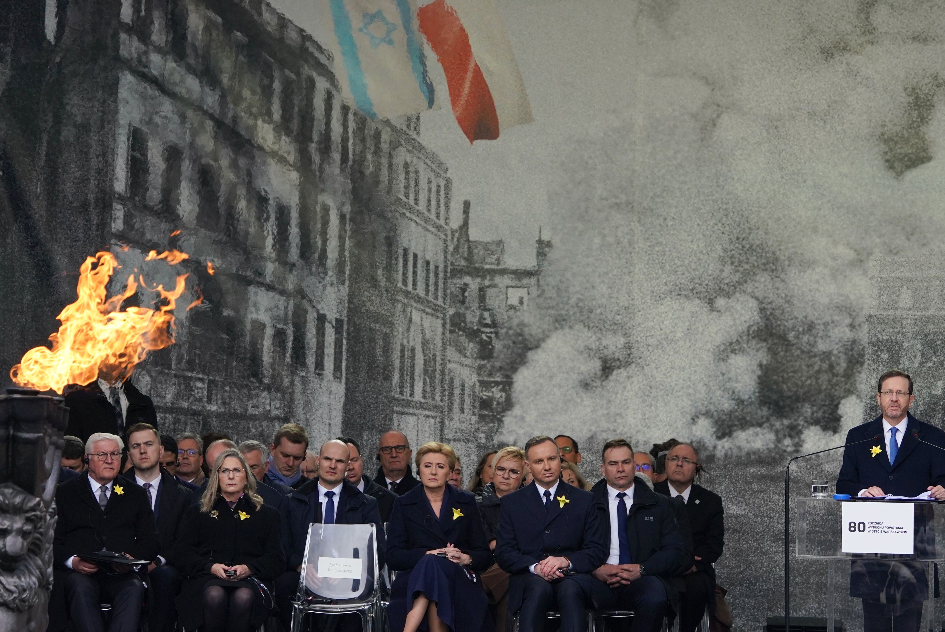 Oficjele siedzą na krzesłach na podeście. Za nimi duże czarno-białe zdjęcie płonącego warszawskiego getta. Na zdjęciu powiewają flagi Izraela i Polski