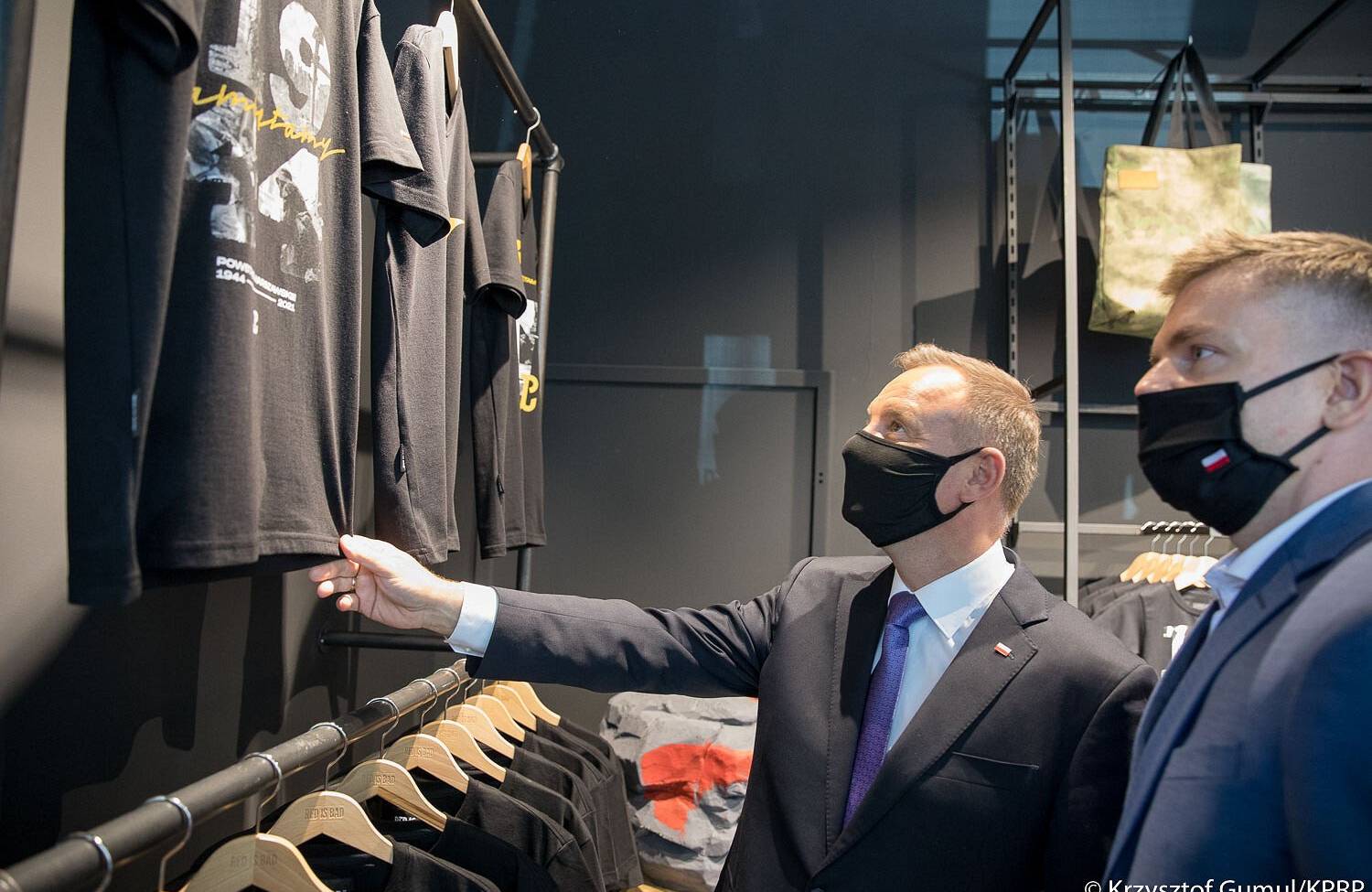 Prezydent Andrzej Duda w towarzystwie szefa firmy Red is Bad Pawła Szopy ogląda odzież w sklepie. Dwóch mężczyzn w czarnych maseczkach i granatowych garniturach stoi przed wywieszonymi na ścianie czarnymi koszulkami. Red is bad