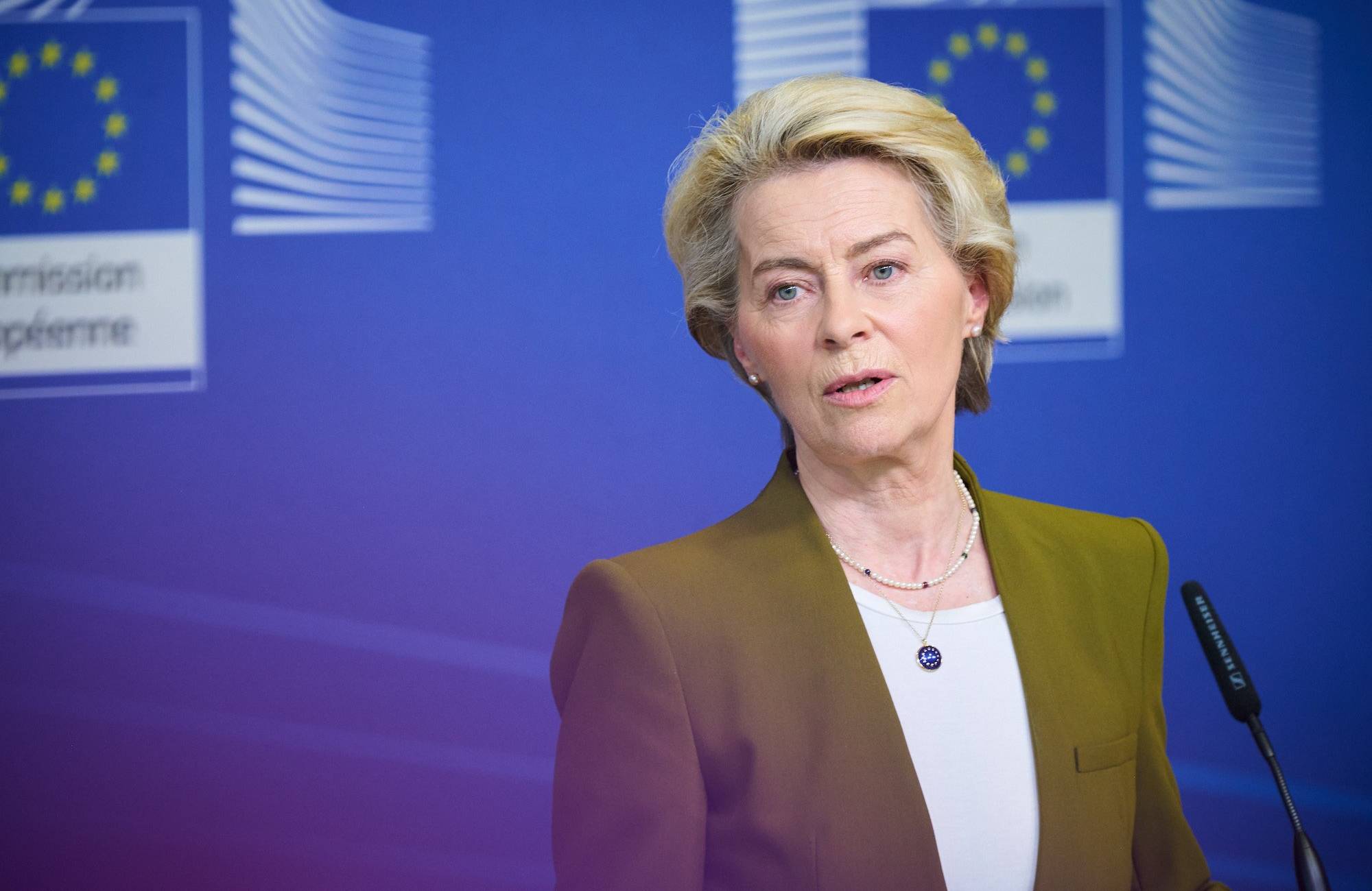 Przewodnicząca Komisji Europejskiej Ursula von der Leyen w oliwkowym żakiecie i białej bluzce, na tle niebieskiej ścianki z logo KE. Przemawia na konferencji prasowej.