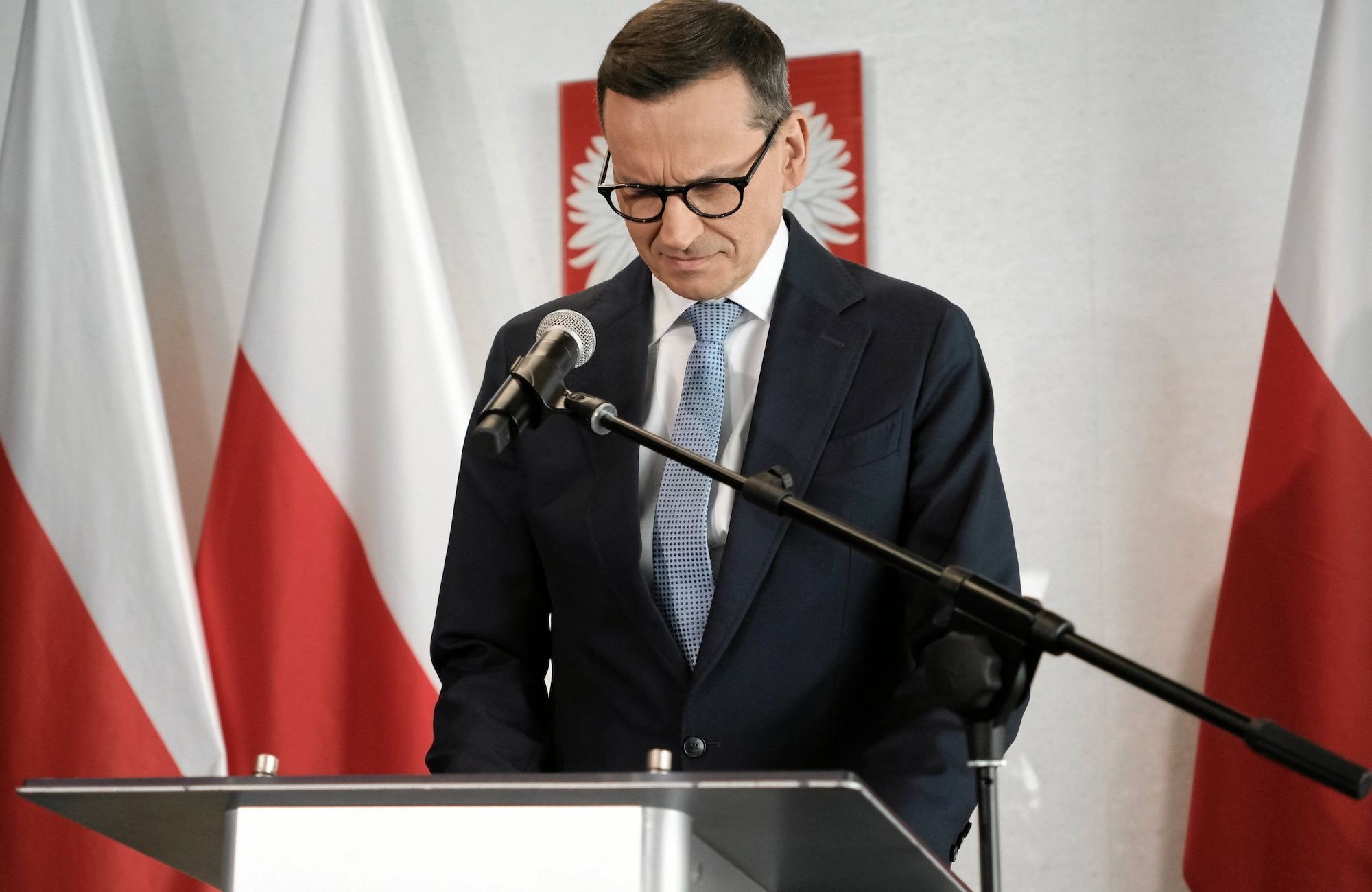 Mateusz Morawiecki przemawia zza podium do mikrofonu na tle biało-czerwonych flag i polskiego godła. Ma spuszczoną głowę