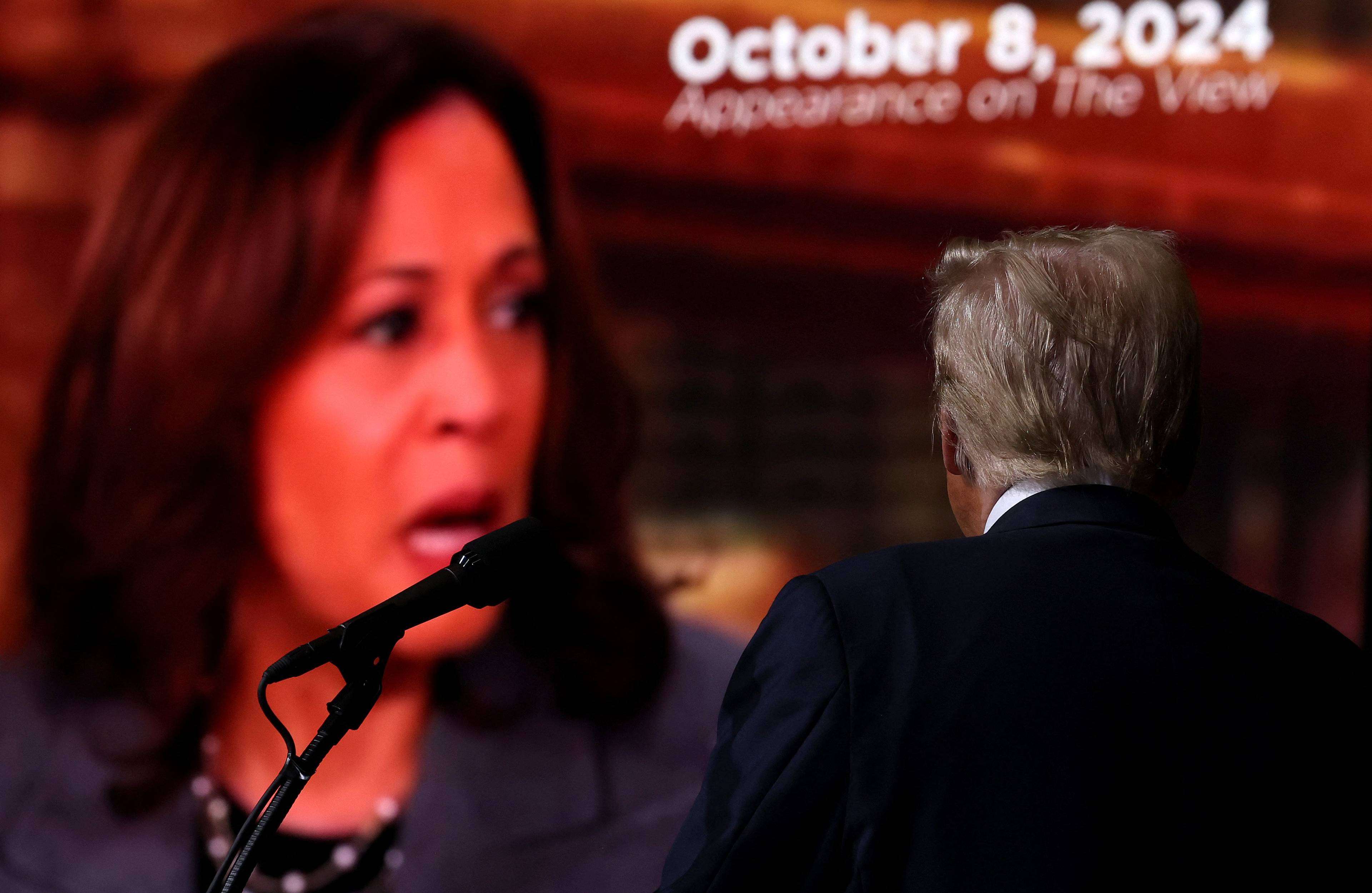 Donald Trump (ujęcie od tyłu) stoi przed wielkim ekranem, na którym widać twarz Kamali Harris