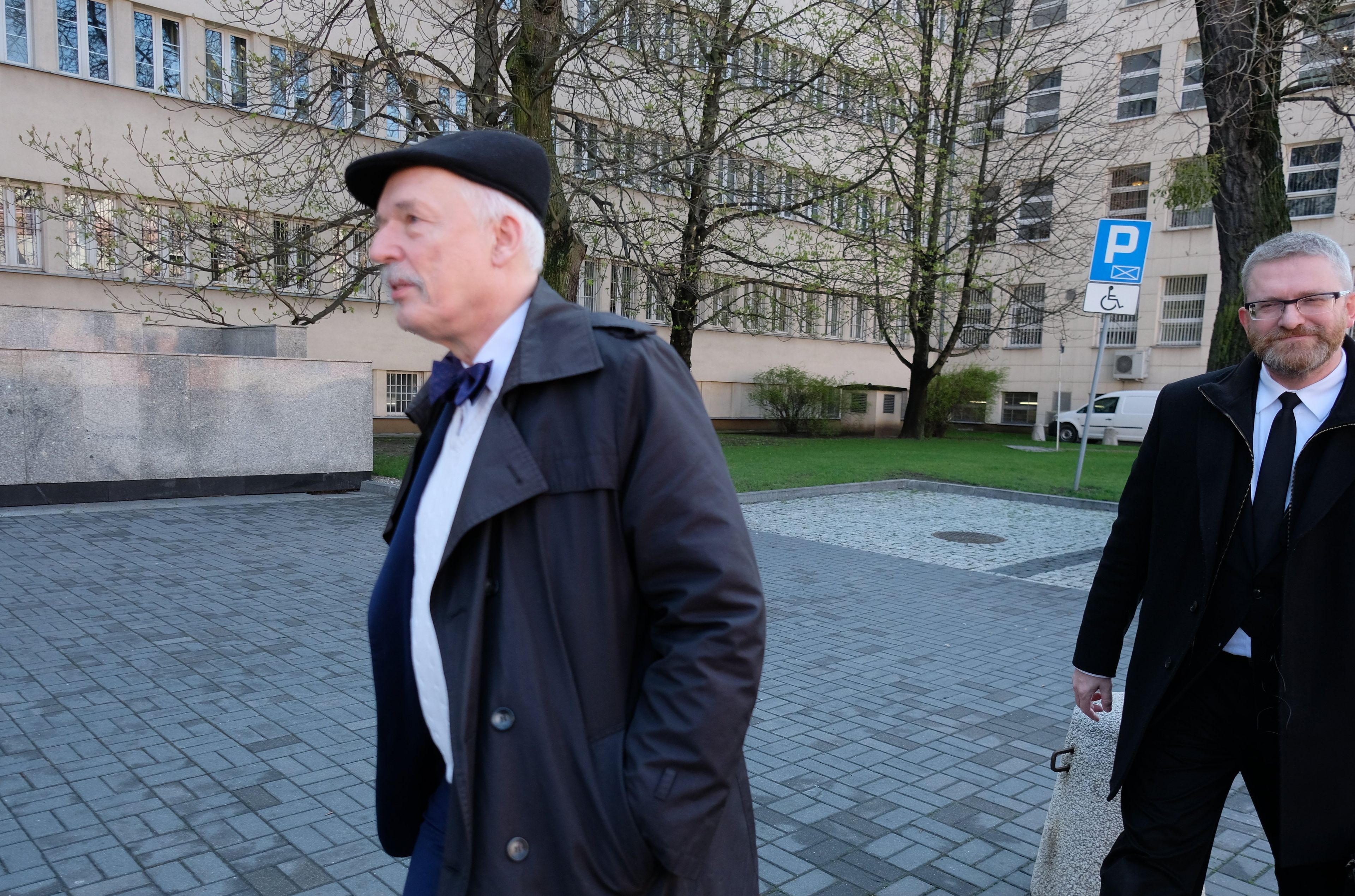 Mężczyzna w kaszkiecie, płaszczu i z muszką pod szyją, za nim mężczyzna z brodą w garniturze i płaszczu