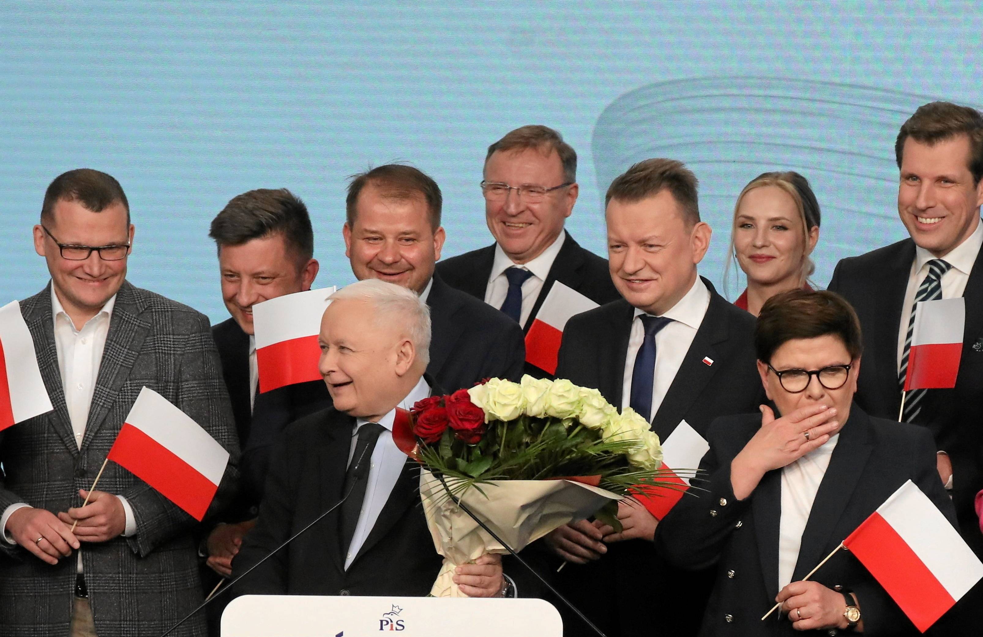 Rozradowany Jarosław Kaczyński - niski siwy starszy pan - w otoczeiu mężczyzn z biało-czerwonymi flagami, z biało-czerwonym bukietem w dłoni