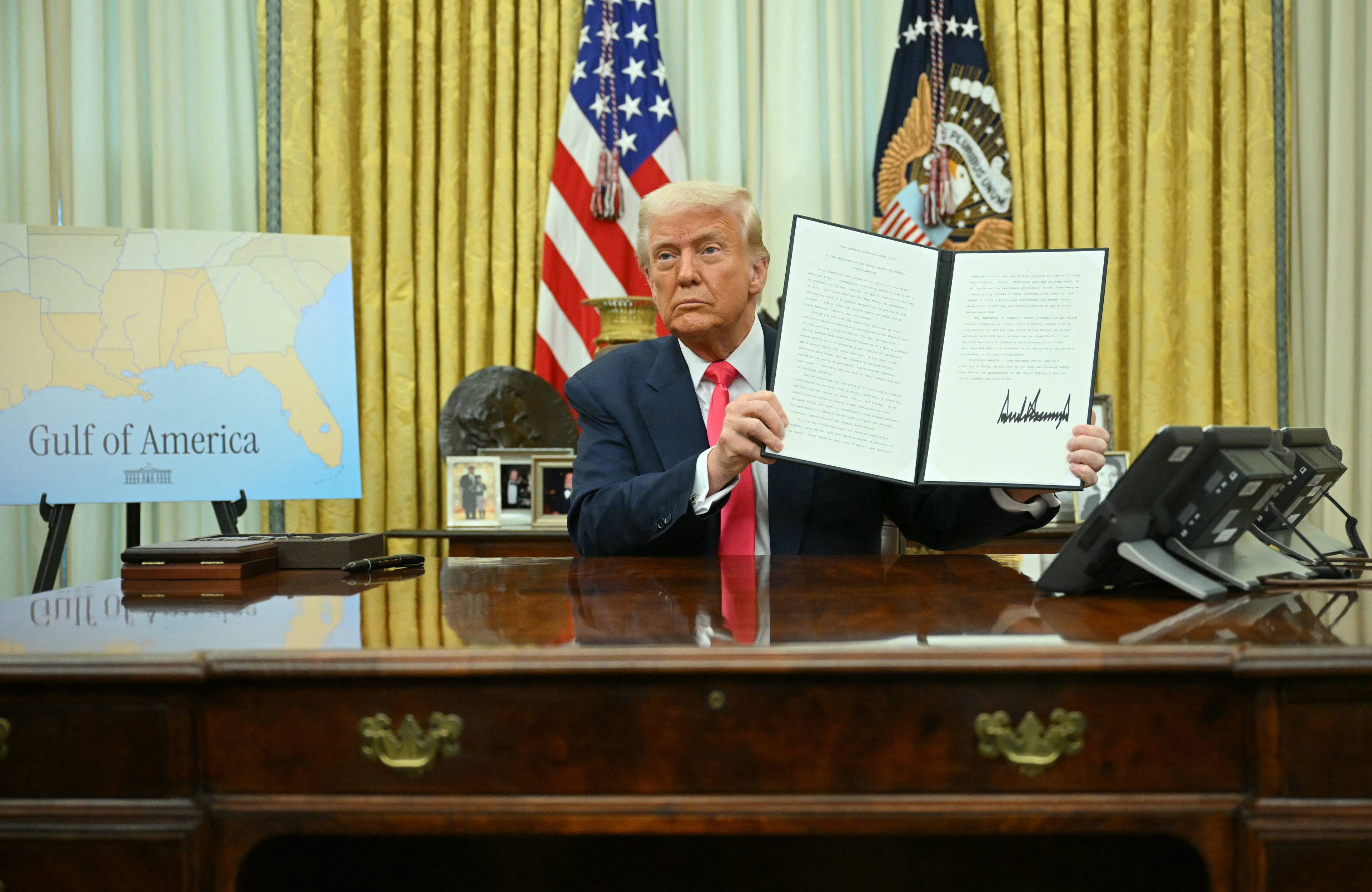 Donald Trump siedzi za biurkiem w Gabinecie Owalnym i pokazuje do kamery dekret ze swoim podpisem