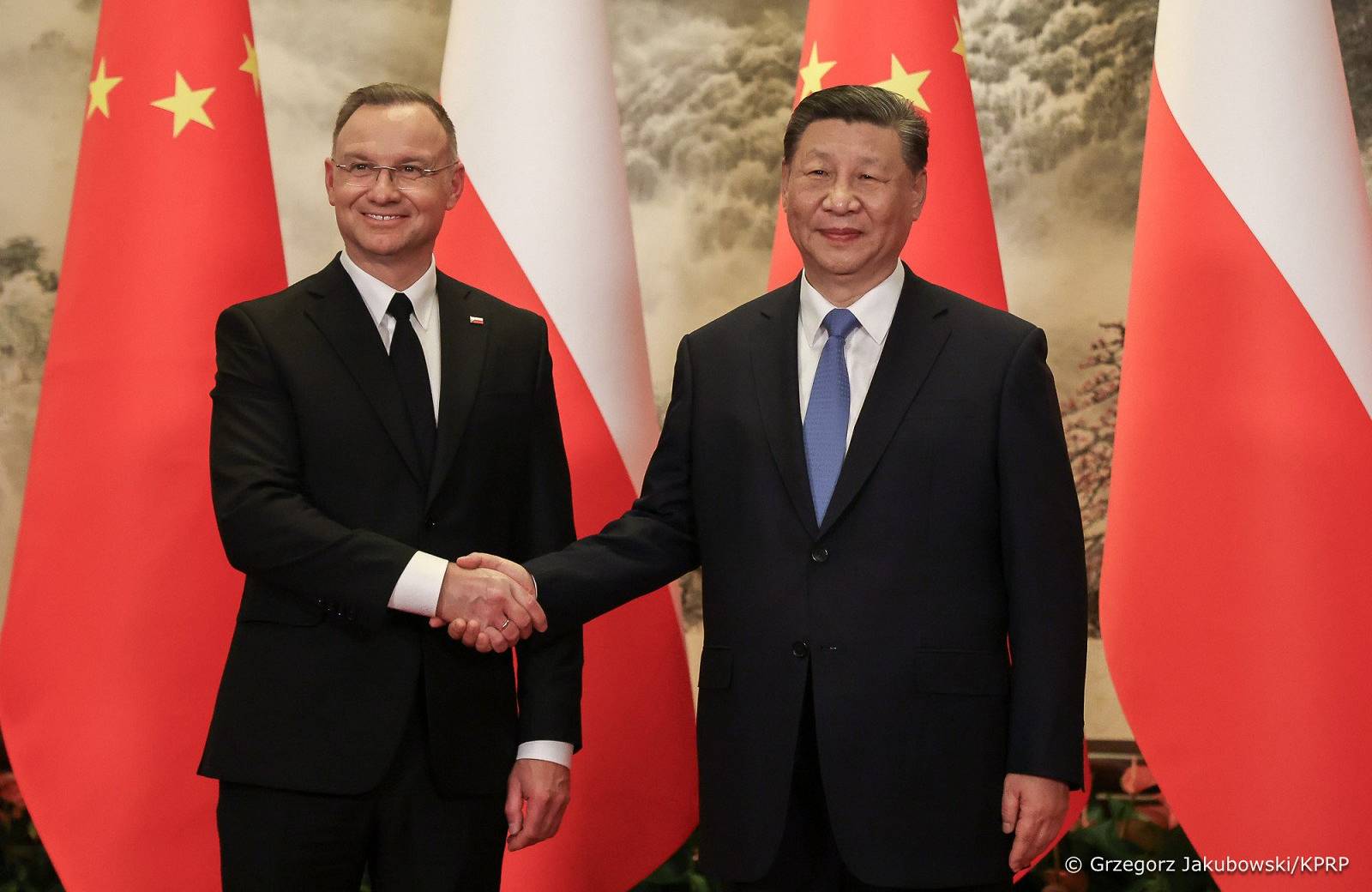 dwaj mężczyźni w garniturach (Duda i Xi) ściskają sobie ręce, w tle flagi