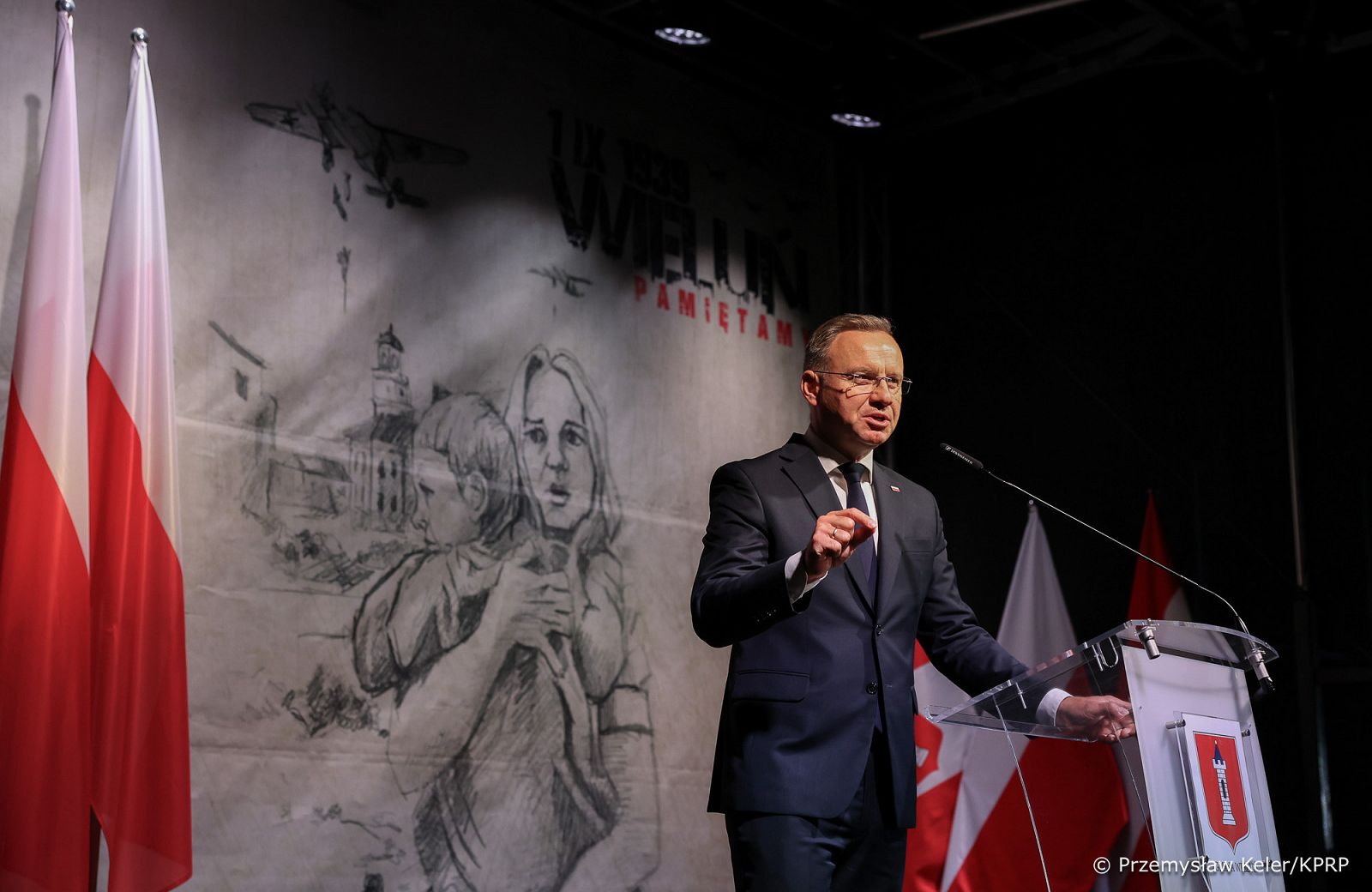 Mężczyzna w czarnym garniturze stoi przy przezroczystej mównicy z mikrofonem, przemawia, w tle rysunek ołówkiem kobiety trzymającej dziecko w ramionach oraz bombardowanego miasta, obok tablicy z rysunkiem polskie flagi.