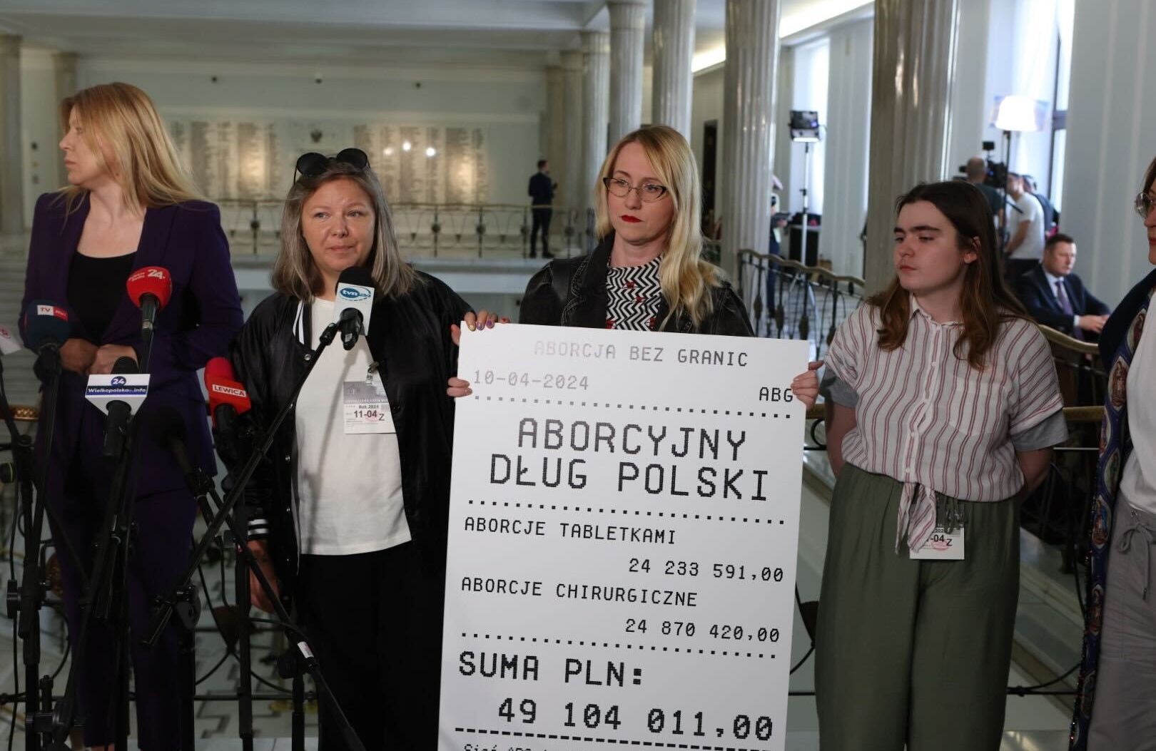 Działaczki Aborcji bez Granic stoją w holu Sejmu podczas konferencji prasowej. Jedna z nich trzyma powiększony paragon z napisem aborcyjny dług Polski