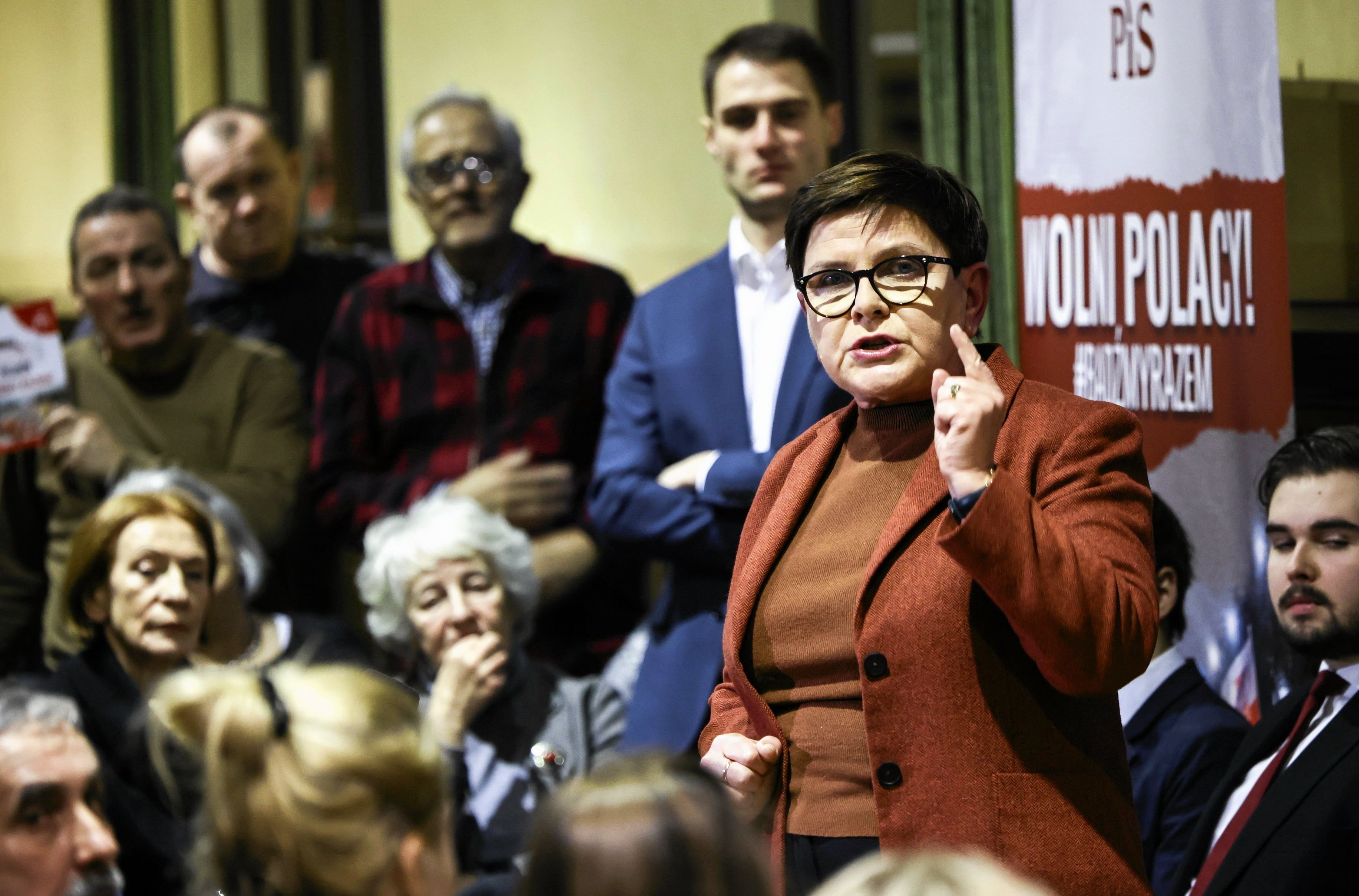 Tłum ludzi siedzi, kobieta w okularach stoi i macha ręką