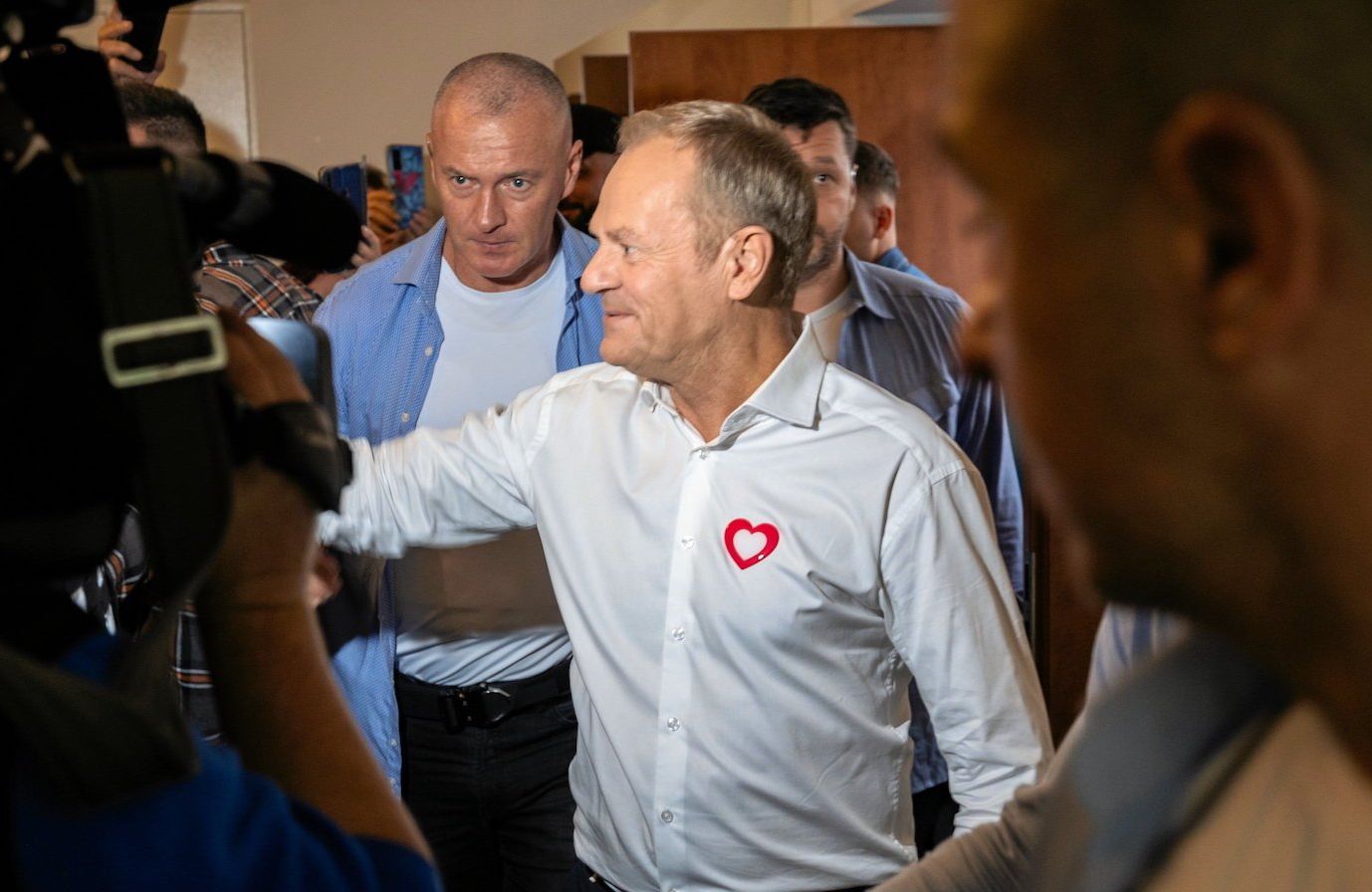 Donald Tusk w otoczeniu reporterów wychodzi z sali i z uśmiechem wyciąga rękę do osoby stojącej po prawej stronie, poza kadrem