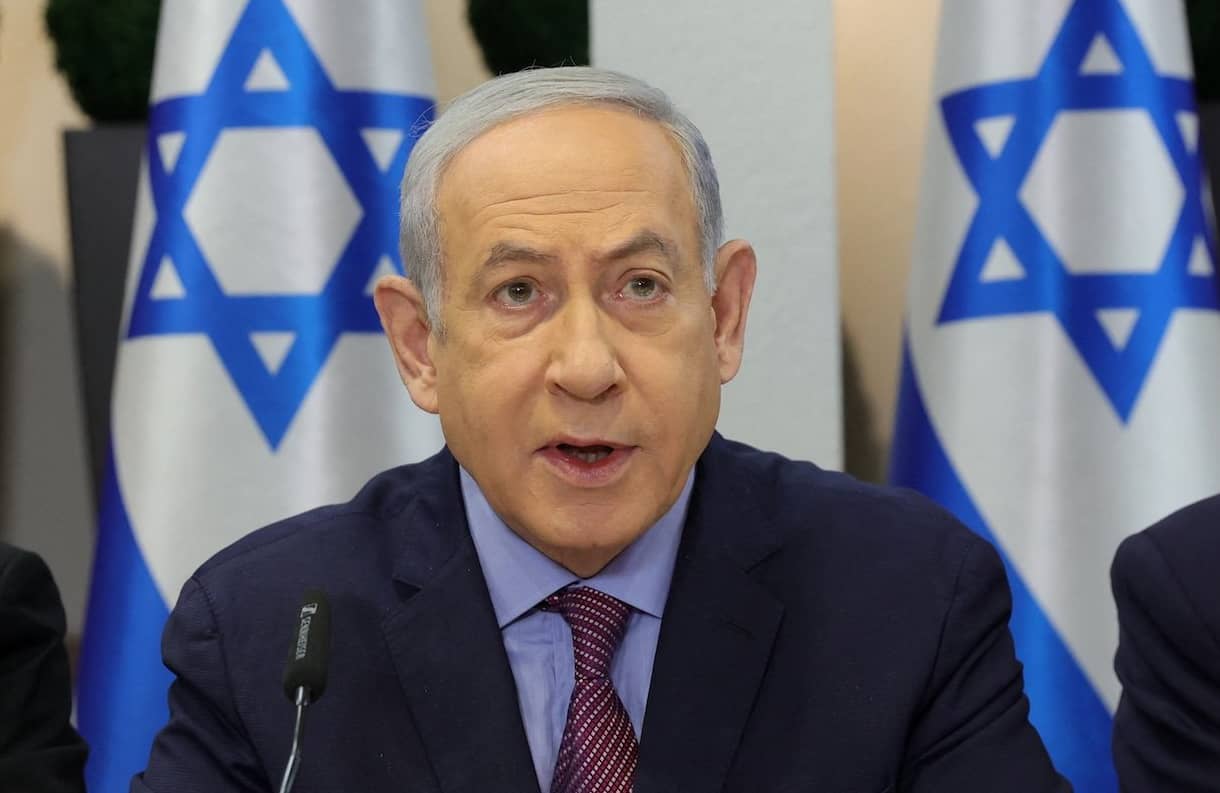 Binjamin Netanjahu przemawia na konferencji na tle flag Izraela. Siedzi za stołem i opiera na nim przedramiona, ma splecione palce dłoni.